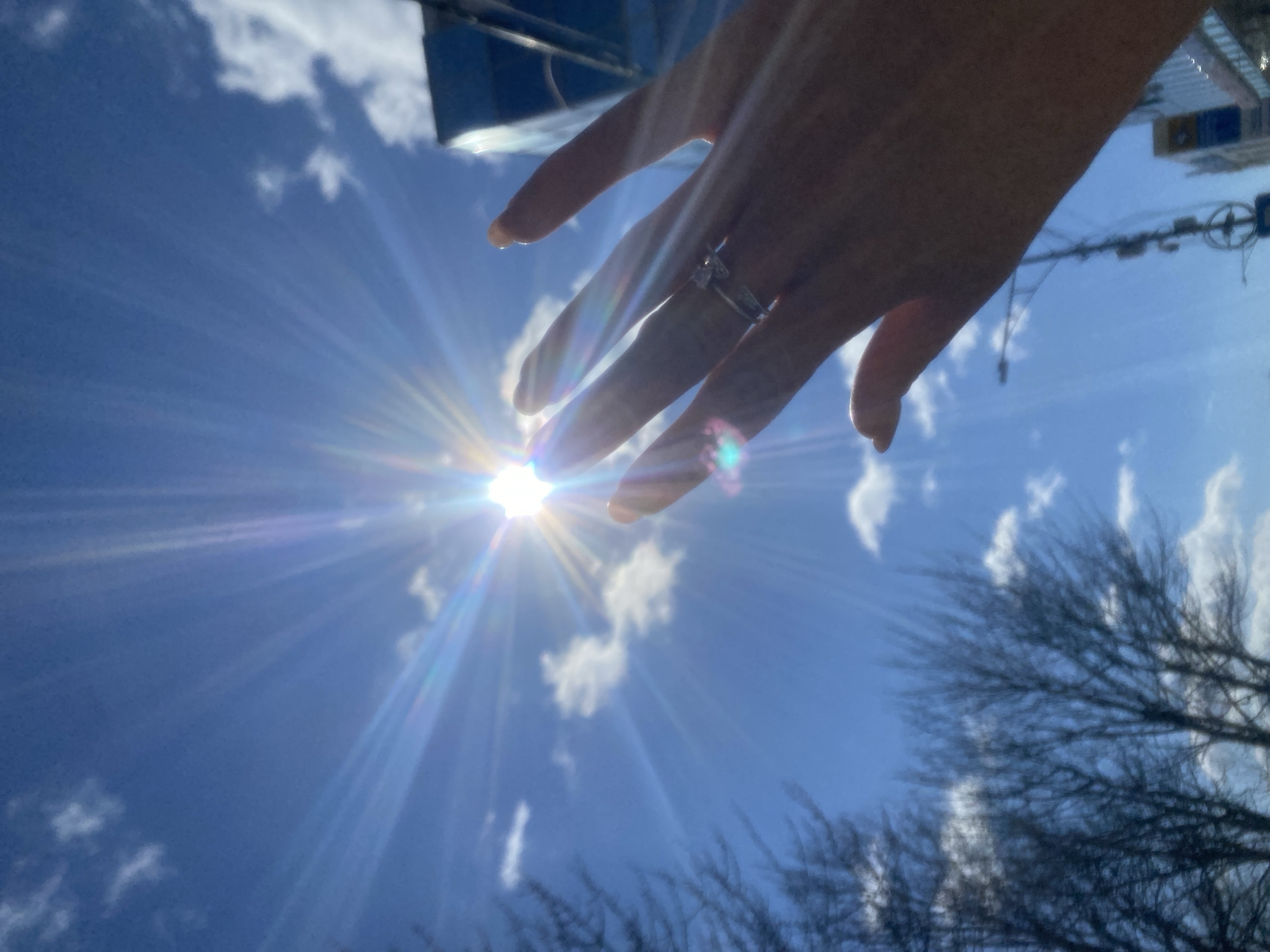 A low-angle shot shows a person's hand reaching toward the sun in a blue sky with scattered clouds. The hand is fair-skinned and has a ring on one finger. The sun is bright and creates lens flare. Bare tree branches are visible along the bottom edge of the image. Reflected upside-down images of buildings and a street light pole can be seen in the background sky above the hand, indicating an urban location. The location is not Petropavlovsk-Kamchatsky as no landmarks are recognizable.