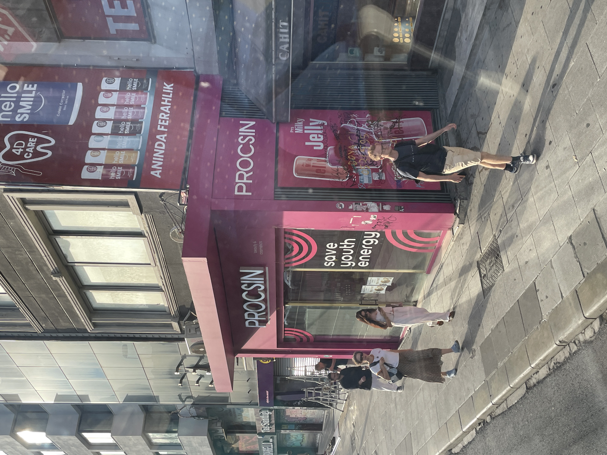 The image shows a street scene in Kadıköy, Turkey. A bright pink storefront, branded "PROCSIN BEAUTY & COSMETICS", is centrally located. Above the entrance, the "PROCSIN" logo is prominently displayed in white lettering. A large advertisement for "hello SMILE" products, featuring toothpaste and dental care items, is visible on the left side of the storefront. To the right of the "PROCSIN" entrance, a dark awning with the name "CAHIT" is visible.
On the sidewalk in front of the storefront, four individuals are present. Two women, one in a white top and patterned skirt, and another in a white outfit, are walking away from the viewer. A man in a dark t-shirt and beige shorts is walking towards the viewer. Another man in a black shirt is standing to the left, near a ladder. The sidewalk is paved with large, square tiles, and a street curb is visible in the foreground. The background consists of multi-story buildings, some with reflective glass facades and architectural details. The sun casts long shadows across the scene, indicating it is likely late afternoon.