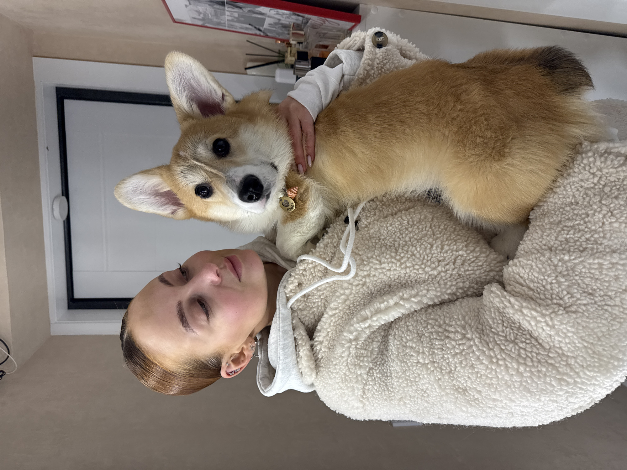A young person with auburn and dark roots, wearing a white fleece hoodie and a long-sleeved white shirt, is holding a Pembroke Welsh Corgi. The dog has a brown and white coat and is wearing a collar with a tag. The person is looking at the camera. A doorframe and a shelf with objects are in the background. The photo was likely taken in Chaikovsky, Russia.