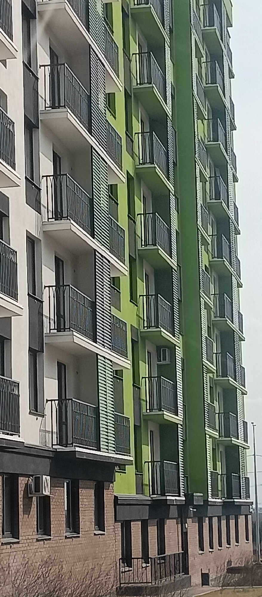 The image shows the exterior of a modern apartment building. The building has multiple stories, each with balconies featuring black metal railings. The building has two sections - one section is painted white and the other a shade of green. The lower portion of the building, visible at the base, appears to be constructed of brick. There is a sidewalk alongside the building. The sky is overcast. Location is likely urban but unknown.