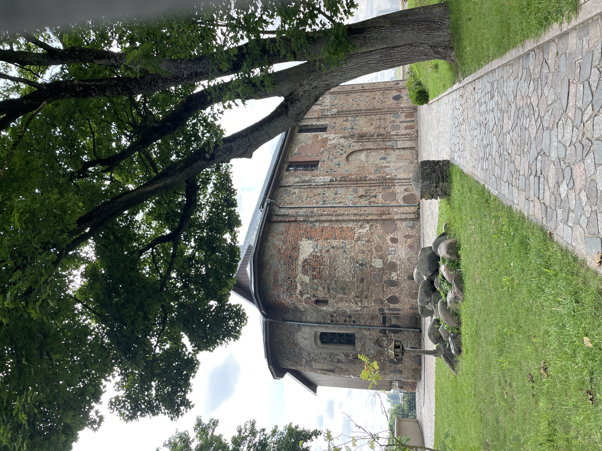 A side view of the Kalozha Church, also known as the Church of St. Peter and St. Paul, in Hrodna, Belarus. The ancient stone and brick structure features a rounded apse and a wooden roof. A large tree with lush green leaves dominates the foreground, casting shadows over a cobblestone pathway. The path leads away from the church, bordered by green grass and some decorative rocks. The sky is partly cloudy, with hints of blue. The image captures a serene and historic atmosphere.