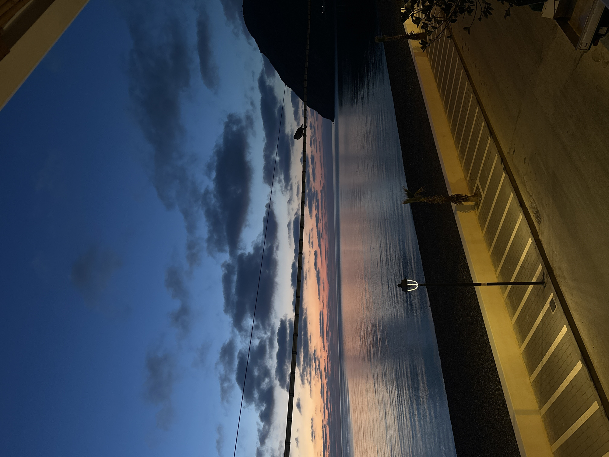 This image captures a tranquil coastal scene at dusk or dawn. The upper portion of the frame is dominated by a sky that transitions from deep indigo blue at the top left to lighter blues and a striking band of orange and pink along the horizon, suggesting a sunset or sunrise. Scattered dark grey clouds are visible against the blue sky.
Below the vibrant horizon, a calm body of water, likely the sea, reflects the sky's colors, appearing dark blue and grey with subtle hints of pink and orange. On the left side, a large, dark, silhouetted mountain or hill rises against the horizon.
A utility pole with a power line stretches horizontally across the middle of the frame, above the water, with a dark bird or object perched on the line.
In the foreground, on the right side, is a paved promenade or street. It features a patterned sidewalk with grey paving blocks and horizontal light yellow or white stripes. A low, light-colored wall or curb separates the sidewalk from a darker, possibly rocky area adjacent to the water. A tall, black streetlamp with a distinctive three-pronged light fixture stands on this paved area. Several small palm trees are visible along the promenade, and darker foliage from bushes or trees is seen in the top right corner. A partial view of a light-colored archway or building overhang frames the top left corner, suggesting the photo was taken from within a covered vantage point.
The scene conveys a peaceful and serene atmosphere, characteristic of a Mediterranean twilight. No discernible text is visible.