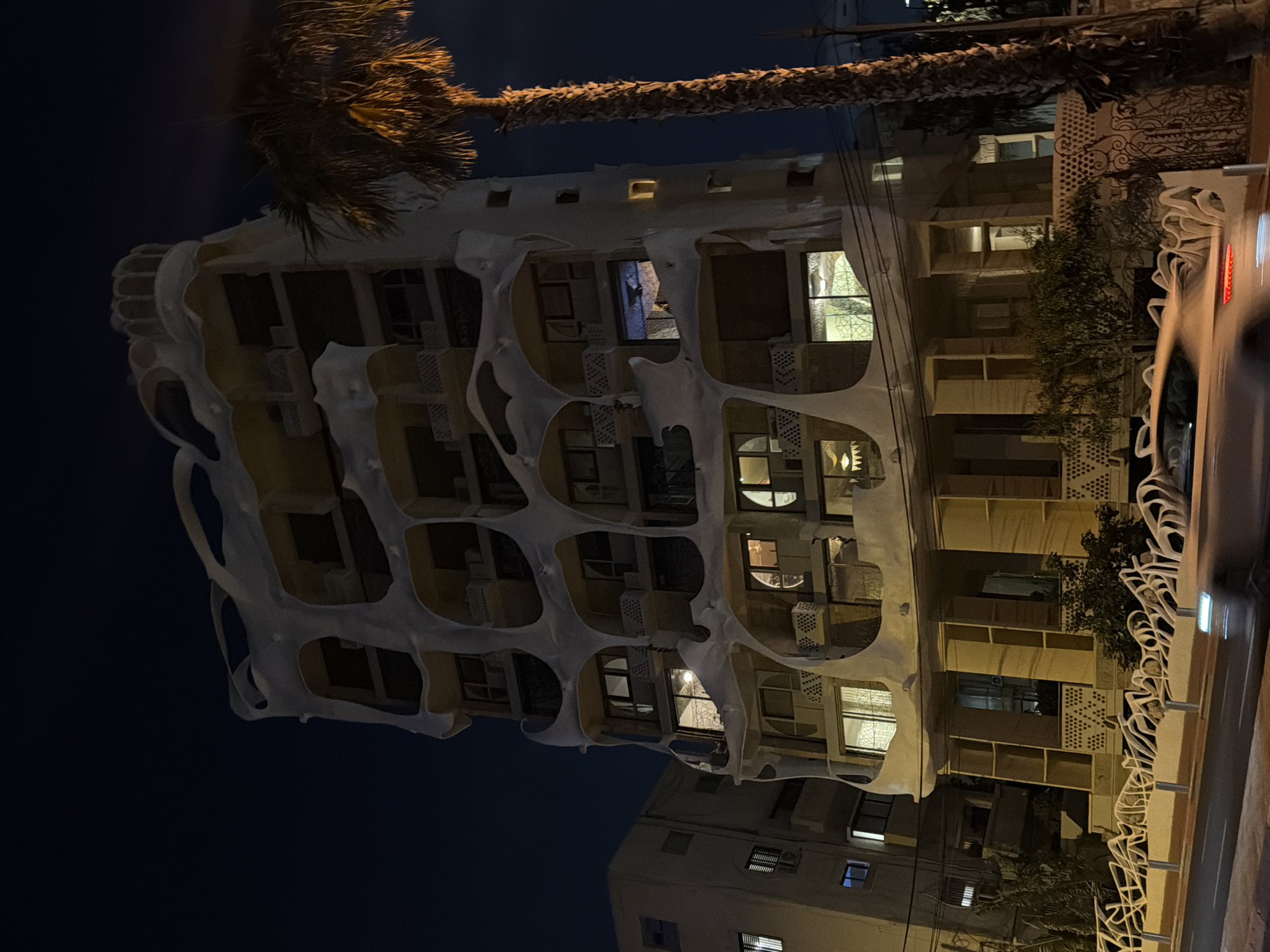 The image shows the exterior of the Gaudi House, a distinctive building located in Tel-Aviv, Israel. It's taken at night. The building is characterized by its white, curvilinear facade reminiscent of the work of architect Antoni Gaudí. Several windows on different floors are illuminated. A palm tree and street lighting are visible, adding to the urban atmosphere. To the right, a car is blurred, suggesting movement. The sky is dark, indicating a late evening or night. The building across the street is visible with its windows lighted as well.