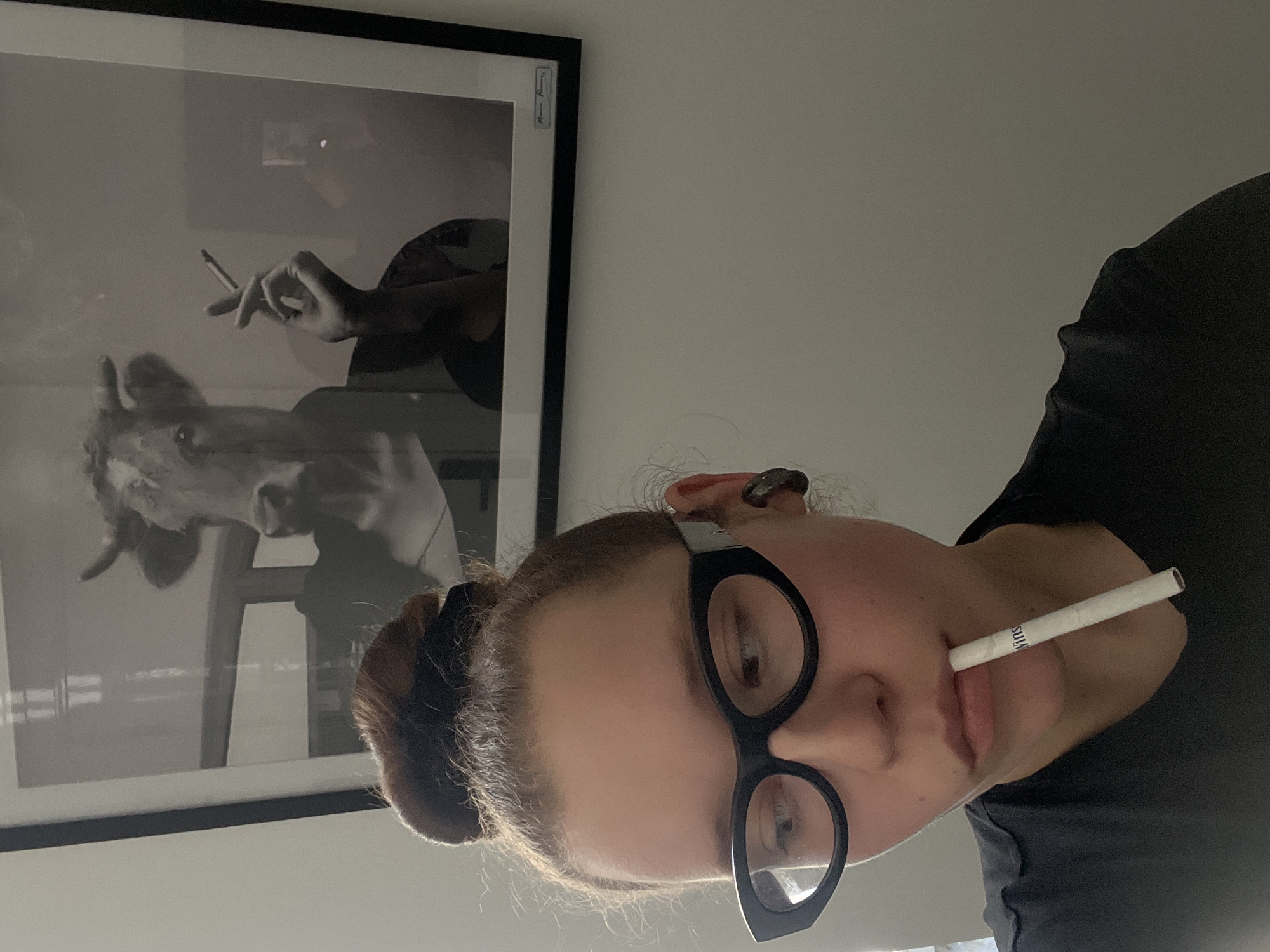 The image depicts a close-up, indoor scene in Tel-Aviv, Israel, focusing on a young individual positioned in the foreground. The subject, seen from the chest up, has light brown hair styled in a messy bun with a dark scrunchie, wears black cat-eye glasses, and a dark t-shirt. A white, unlit cigarette with "Winstons" visible on its side is held horizontally between their lips. A dark earbud or hearing device is present in their right ear. The subject's gaze is directed downwards and to the right.
In the background, mounted on a plain, light-colored wall, is a framed black and white photograph. This artwork portrays a cow's head with horns, superimposed on a human-like torso dressed in a dark garment. The cow's right hand, depicted as a human hand, holds a lit cigarette from which smoke rises. Reflections, suggestive of an indoor light source or window, are visible on the photograph's glass frame. A signature or inscription is present on the lower right matting of the framed artwork.