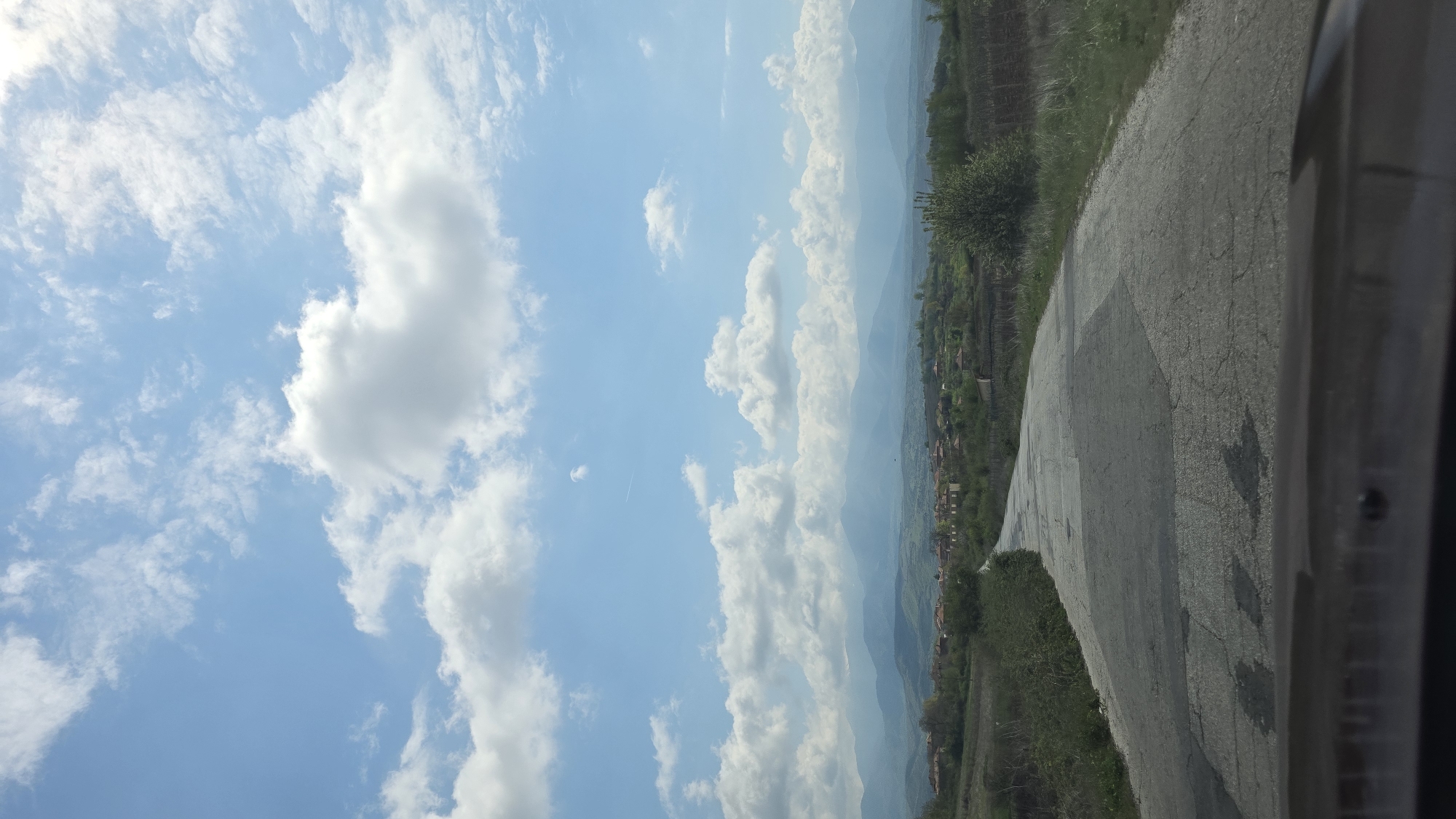 A low-angle shot displays a weathered asphalt road curving to the right, bordered by green vegetation on the far side. Beyond the vegetation lies a landscape with houses and, in the distance, a mountain range obscured by atmospheric haze under a blue sky scattered with white clouds. The perspective suggests the photo was taken from inside a vehicle, as indicated by a partially visible object in the bottom right corner. This scene is located in Harsovo, Bulgaria.