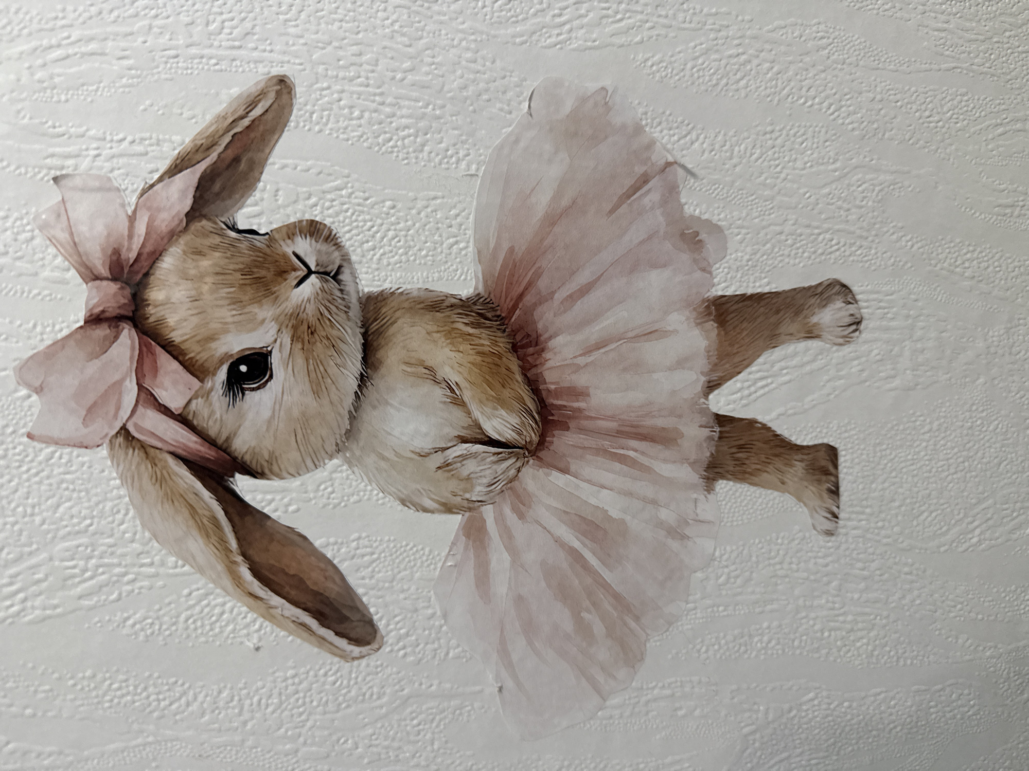 This is a close-up, top-down, eye-level shot of a watercolor illustration of a bunny wearing a pink tutu and a pink bow on its head. The bunny is depicted lying on its back, with its ears spread out and its body bent to the side. Its paws are clasped together in front of its chest. The bunny has large, dark eyes and a small, closed mouth, giving it a serene or content expression. The tutu is made of delicate, layered pink fabric, and the bow is also a soft pink. The background is a textured, light-colored surface, possibly wallpaper, with an embossed pattern of horizontal wavy lines and small bumps. The lighting is even, suggesting it is an indoor setting or a well-lit environment. There is no visible text in the image. The style of the illustration is soft and whimsical, with watercolor washes creating a gentle, ethereal feel.
