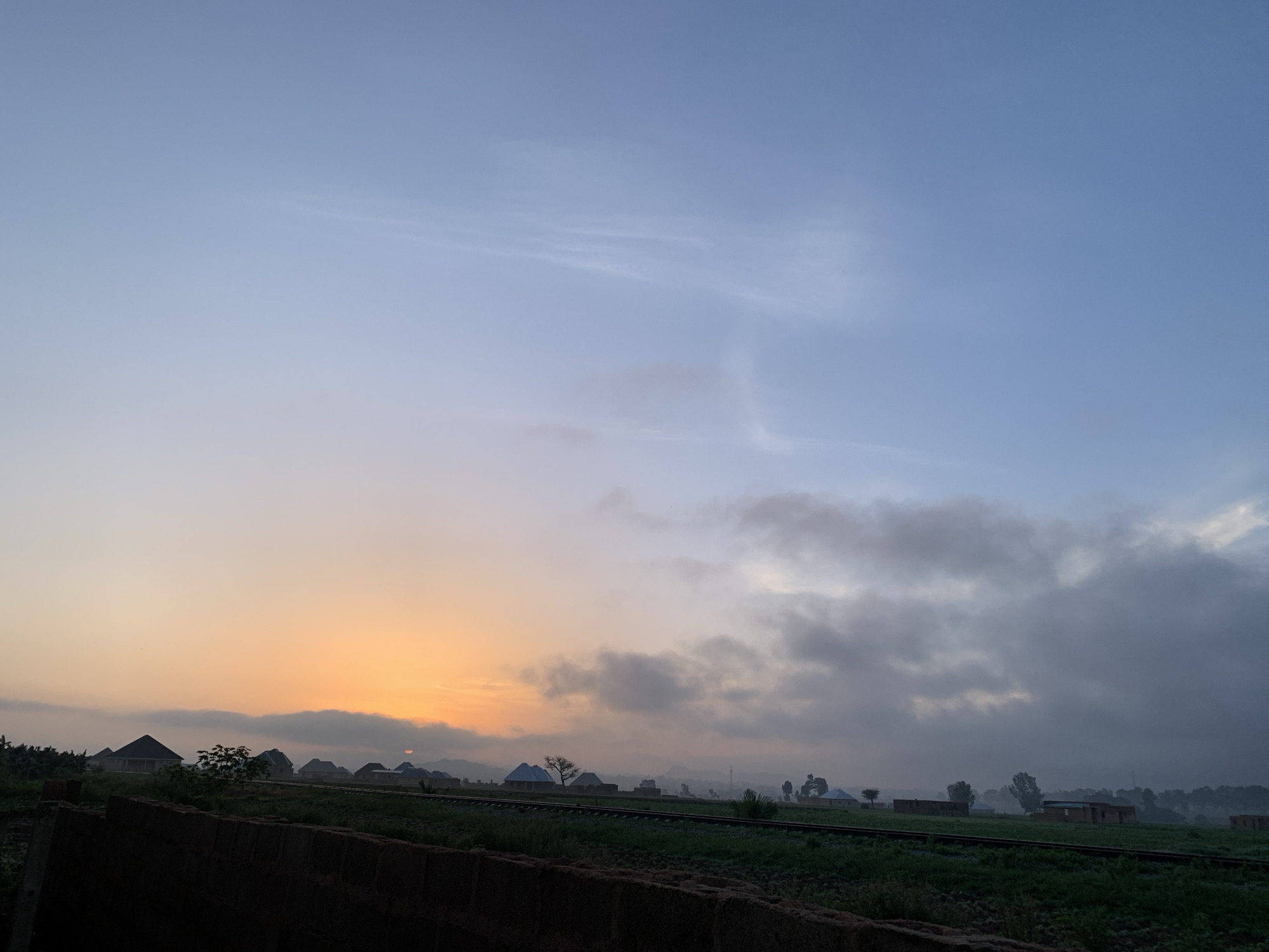 The image captures a sunrise over Kuru Karama, Nigeria. The sky transitions from a soft orange glow near the horizon, where the sun is partially obscured by clouds, to a pale blue overhead. Wispy clouds are scattered across the sky, with denser, darker clouds concentrated towards the right side of the frame. In the middle ground, a line of houses with distinct architectural styles is visible, nestled amidst greenery. Some of the houses appear to be under construction, with exposed brickwork and unfinished roofs. A set of railway tracks runs horizontally across the scene, bordered by green fields. In the foreground, a dark, textured wall, possibly made of brick or stone, runs along the bottom of the image, partially obscuring the view of the landscape. The overall atmosphere is one of a tranquil early morning in a developing area.