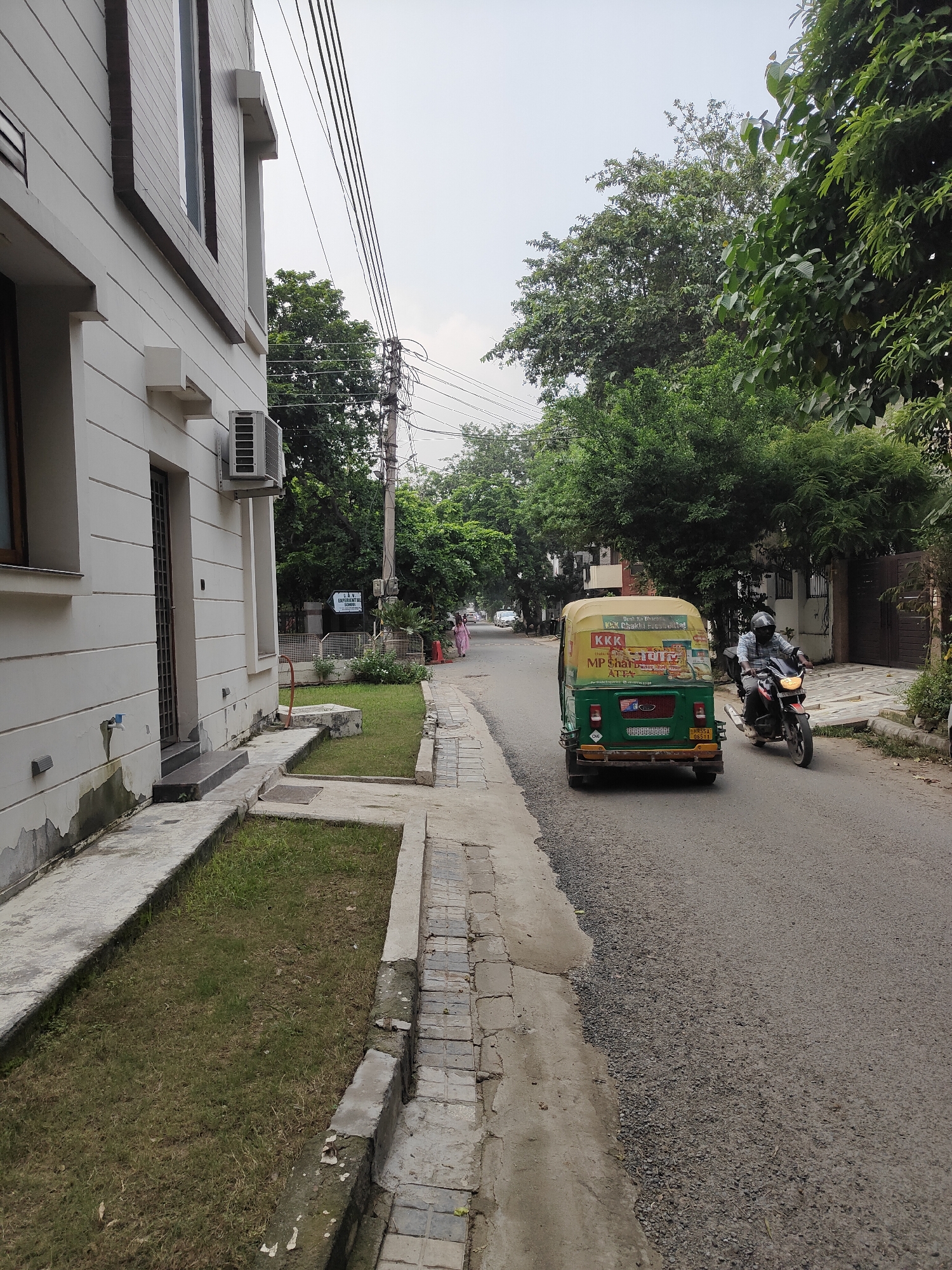 This is a medium-shot, eye-level, vertical, outdoor photograph captured on a cloudy day. The scene depicts a residential street in Gurgaon, India. On the left, a modern, light-colored building with horizontal siding and dark trim dominates the foreground. An air conditioning unit is mounted on the exterior wall. Adjacent to the building, a concrete pathway runs alongside a narrow strip of grass.
The street itself is paved with asphalt and appears to be under some repair with loose gravel along the edge. A green and yellow auto-rickshaw is moving away from the camera, displaying advertisements on its rear. To the right of the auto-rickshaw, a man wearing a helmet and a plaid shirt is riding a motorcycle, also moving away from the camera. Further down the street, trees line the right side of the road, and a fence encloses a property. Two women are walking away from the camera on the sidewalk in the distance. A sign that reads "IAW EXPERIMENTAL SCHOOL" is visible mounted on a pole near the trees. The overall impression is of a quiet, residential neighborhood in a city.