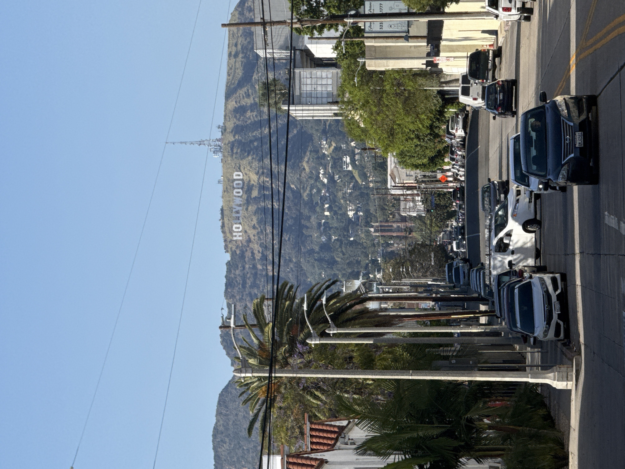 The image depicts a street in Los Angeles, United States, lined with parked and moving cars. The iconic Hollywood sign is visible in the distance on a hillside. Palm trees and other vegetation are interspersed along the street. Utility poles with wires run alongside the road. The sky is clear and blue. The angle of the shot suggests the picture was taken from a medium height.