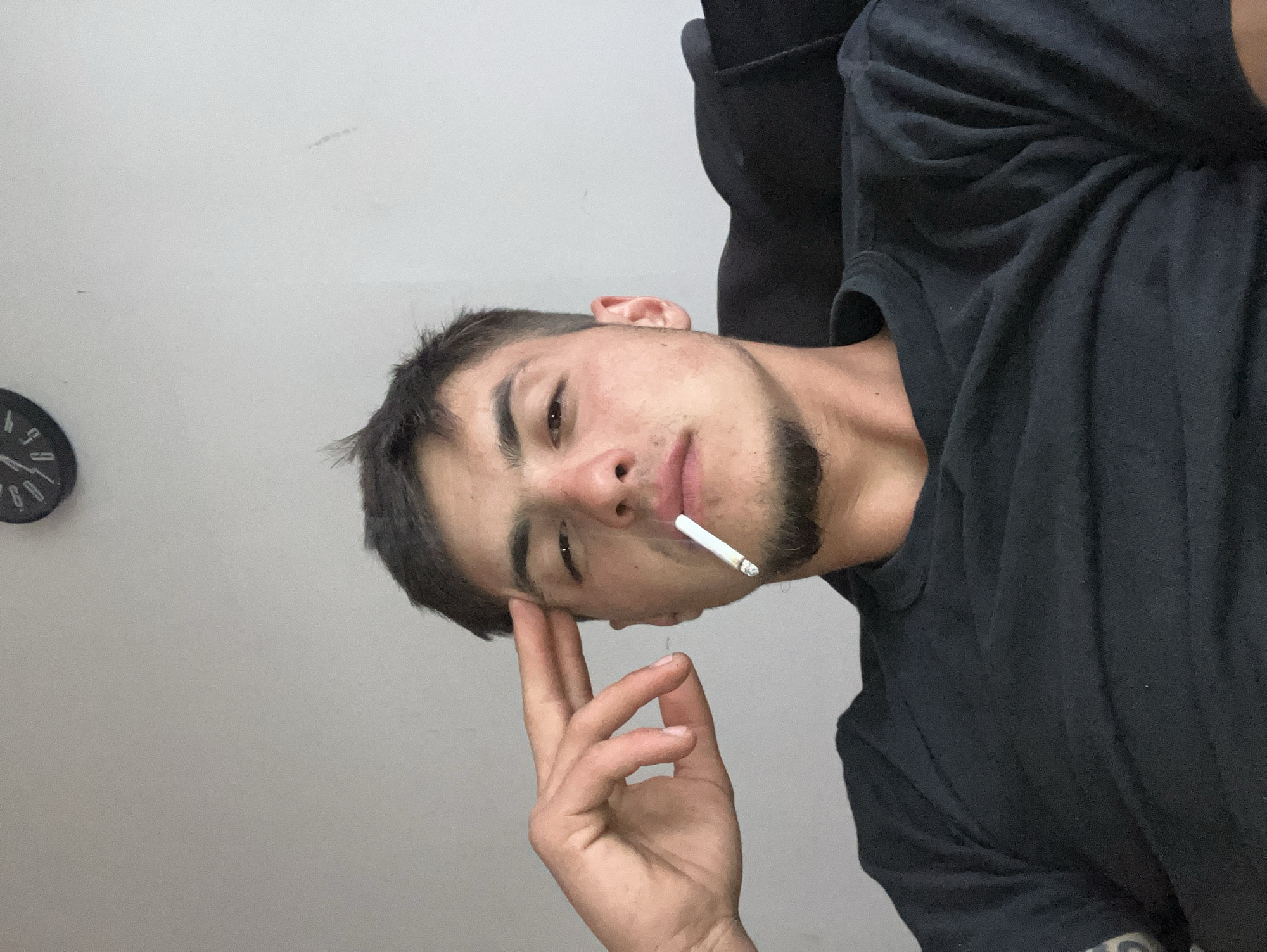 A young adult male is centered in the frame. He is fair-skinned with dark hair and a short beard. He is wearing a black t-shirt. He is holding a lit cigarette in his mouth and his right hand is raised to his forehead with his index finger touching his temple. The background is a plain white wall. A clock is visible in the upper left corner. The location is not apparent from the details in the image. The city of Plovdiv, Bulgaria, cannot be verified.