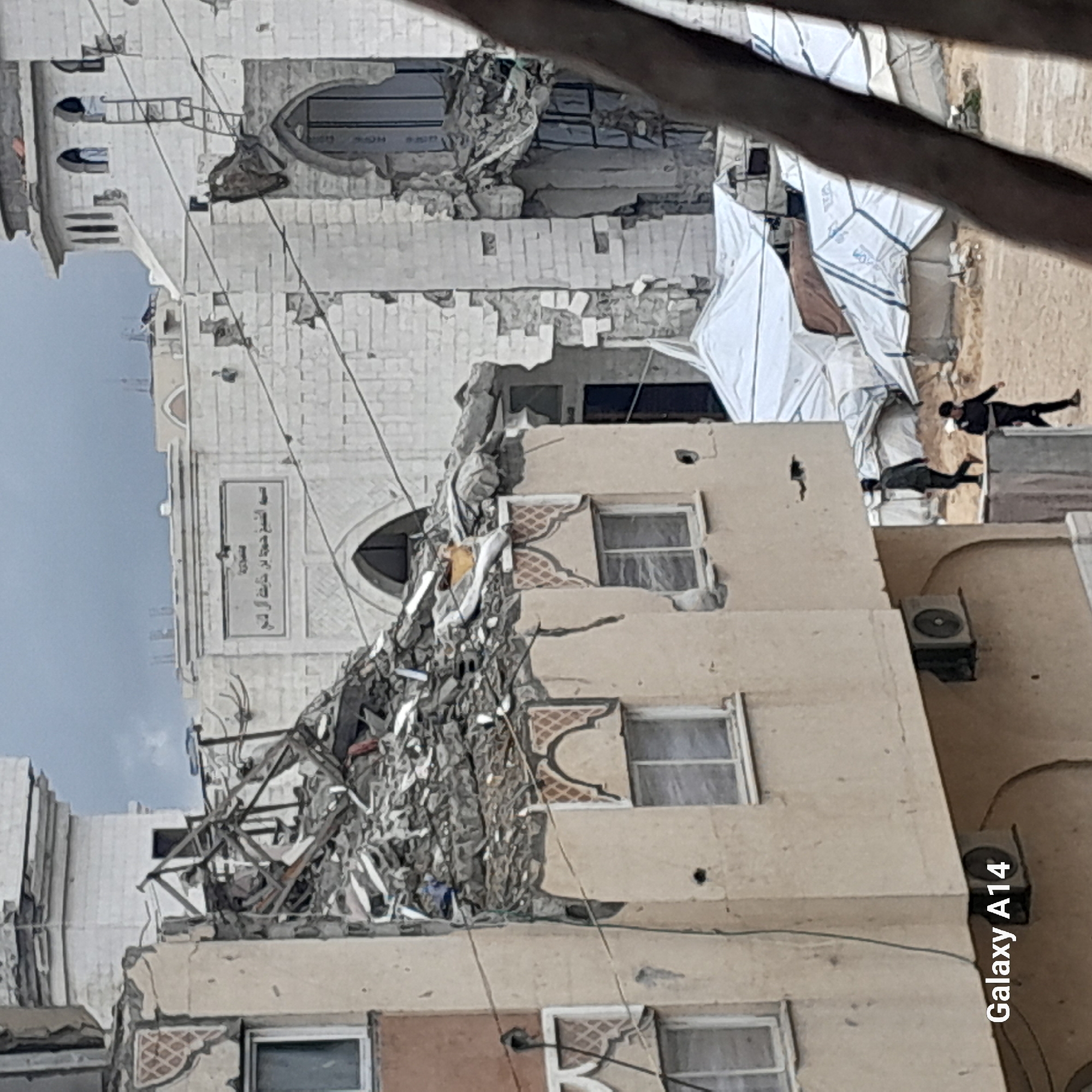 The image depicts a damaged building in Khan Yunis, Palestinian Territory. The building has sustained significant structural damage, with debris visible on the facade. A mosque is partially visible in the background. Two people are seen lying on the ground near what appears to be temporary shelter. An "Galaxy A14" watermark is present in the lower right corner.