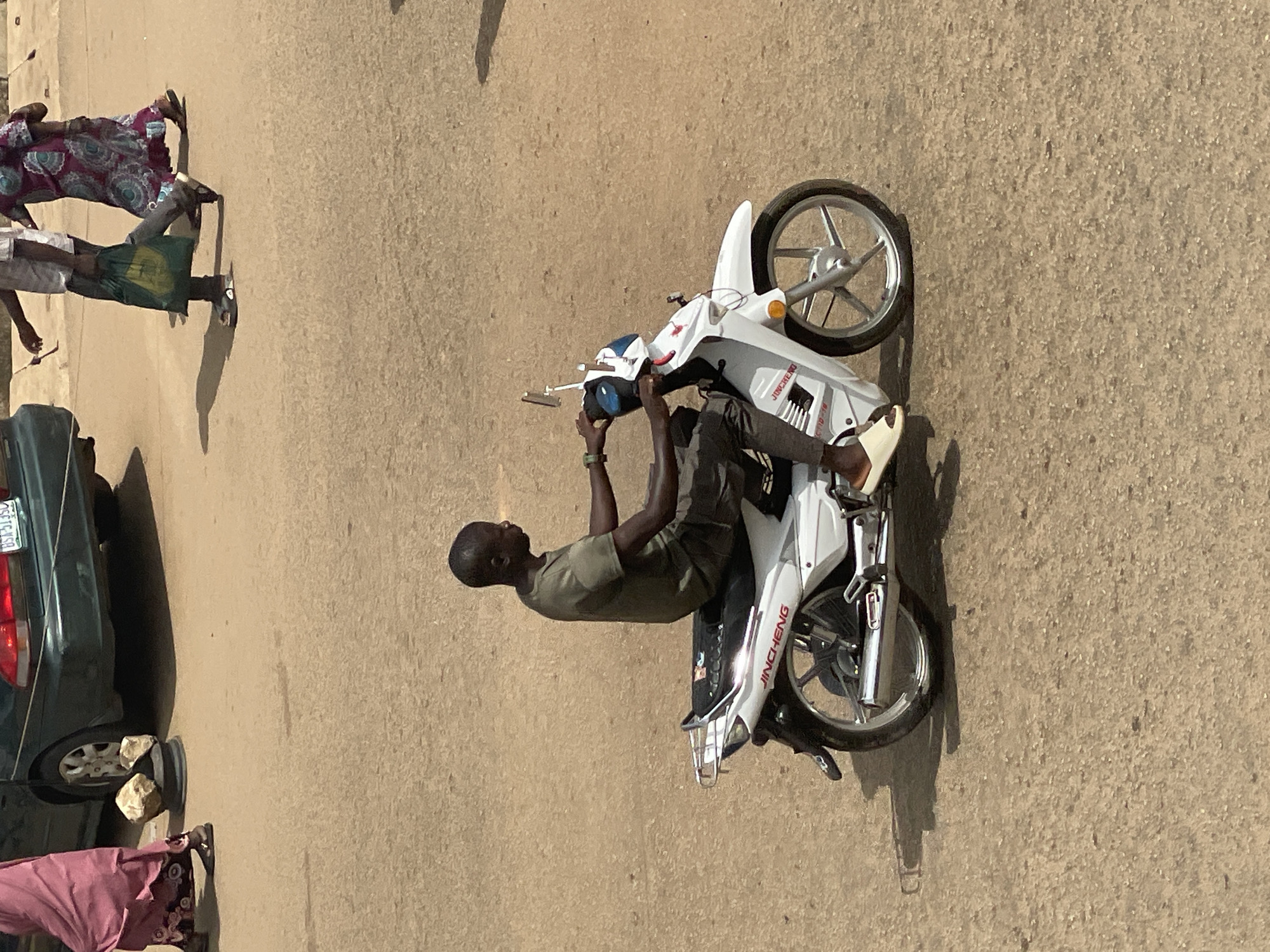 The image shows a man sitting on a white motorcycle on a sandy or dirt road. The man is wearing a green t-shirt and dark pants. He is holding the handlebars of the motorcycle, and appears to be either waiting or preparing to move. To the left of the motorcycle, a dark green car is parked, and there are a few people in the background wearing traditional clothing. The scene appears to be outdoors during the daytime, with the sun casting shadows. The overall atmosphere seems casual, possibly indicating a street scene in a town or village. There is text on the side of the motorcycle that reads "JINCHENG". The image is oriented vertically, with the subjects appearing sideways.