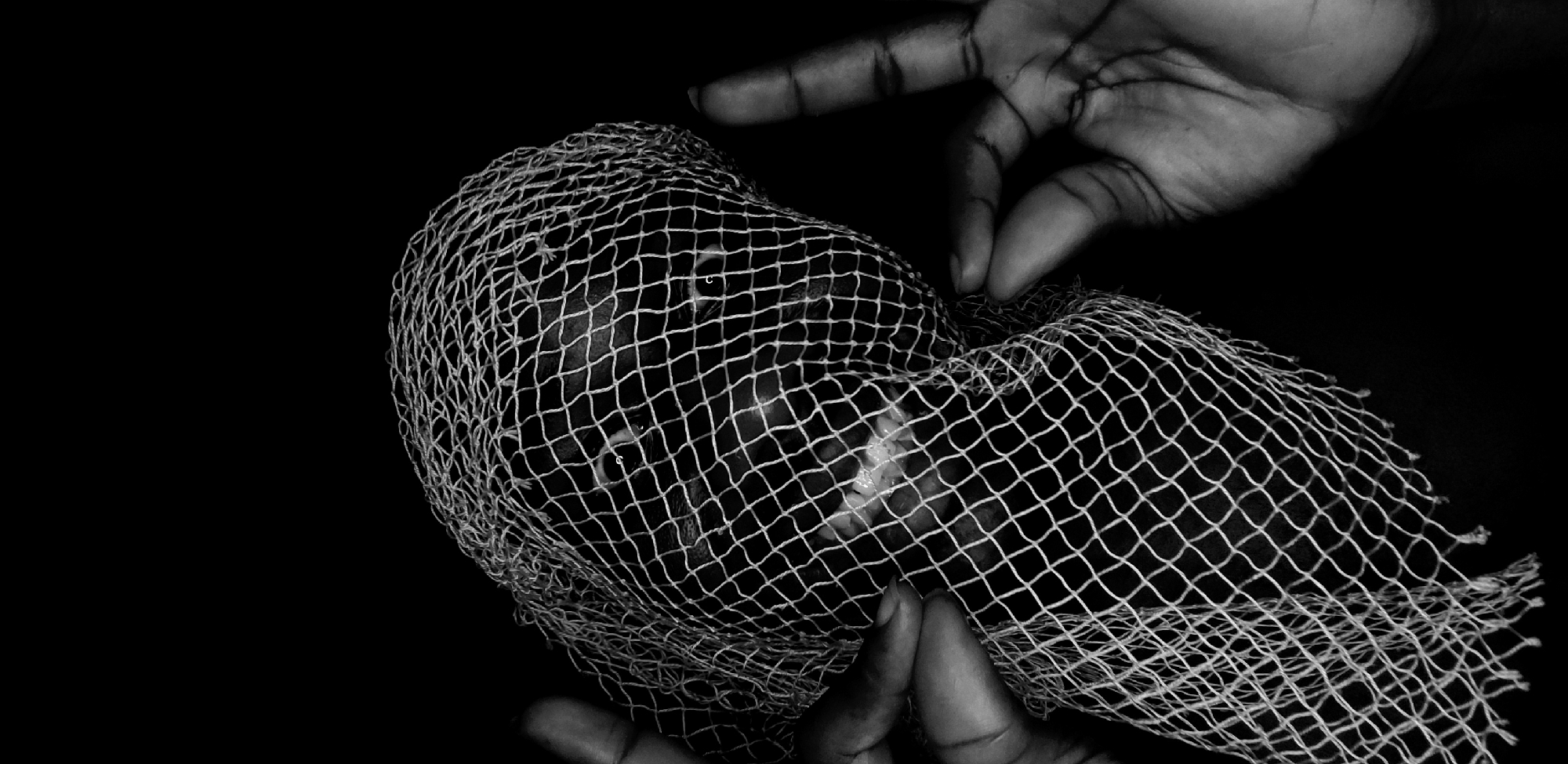 The image is a black and white photograph showing a person's head encased in a white mesh net. Two hands are visible; one appears to be holding the net at the bottom, while the other is positioned near the top of the head. The person's face is visible through the mesh, including the eyes and mouth. The background is entirely black. There is no additional information to confirm the location in Narayi, Nigeria.