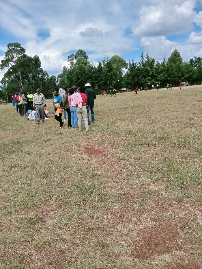 The scene depicts an outdoor grassy field in Lwandeti ward, Kenya, under a partially cloudy blue sky. In the mid-ground, a group of approximately 15-20 individuals, appearing as adults and young adults in casual attire, are gathered, predominantly facing towards the right, observing an activity. Some light-colored items, possibly bags or clothing, are visible on the ground near their feet on the left. Further in the background, a football (soccer) match is in progress on the same field, involving at least 8-10 players wearing red and green/light green jerseys. A white football is visible in mid-air, and goalposts are present on the right side of the playing area. The field surface consists of dry, short grass with visible patches of exposed soil. The background features tall, broadleaf trees on the far left and a dense line of coniferous trees bordering the playing field across the right and center.