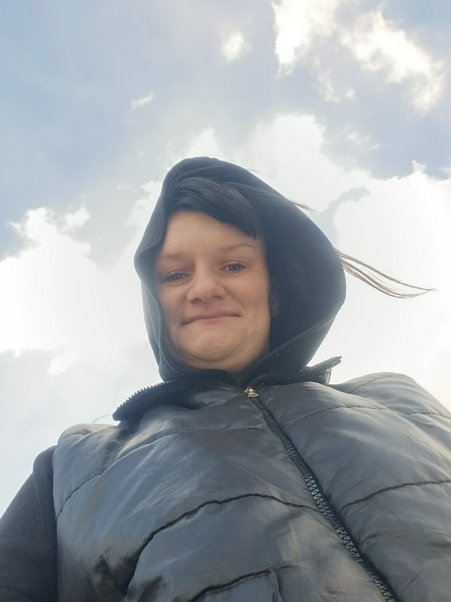 A low-angle, medium shot depicts a woman wearing a hooded black vest against a cloudy sky. The woman's face is visible within the hood, and a few strands of hair are blowing in the wind. The vest is zipped up, and the zipper is visible. The sky is filled with various cloud formations. There is no indication of Фоки, Russia, or any city in the image.