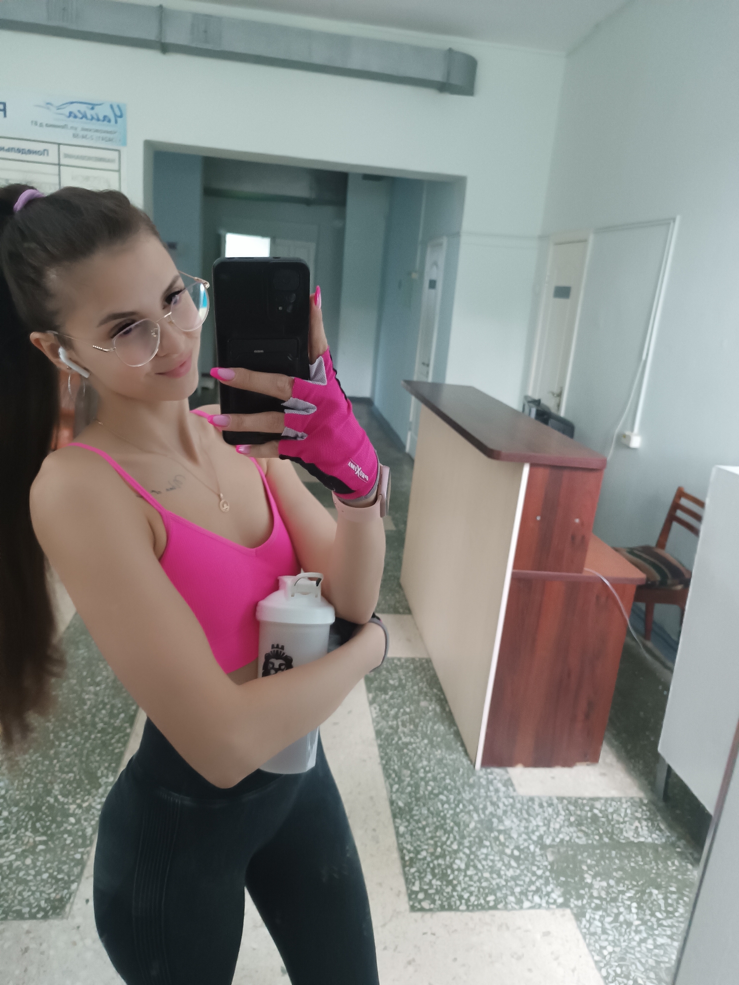 A young woman with long dark hair pulled into a ponytail and wearing glasses and a pink sports bra is taking a selfie in a gym. She is holding a white shaker bottle in her left hand and wearing pink workout gloves. She is wearing black leggings. In the background, there is a reception desk and a chair. The floor is tiled with a speckled pattern. The lighting is bright, suggesting an indoor setting.