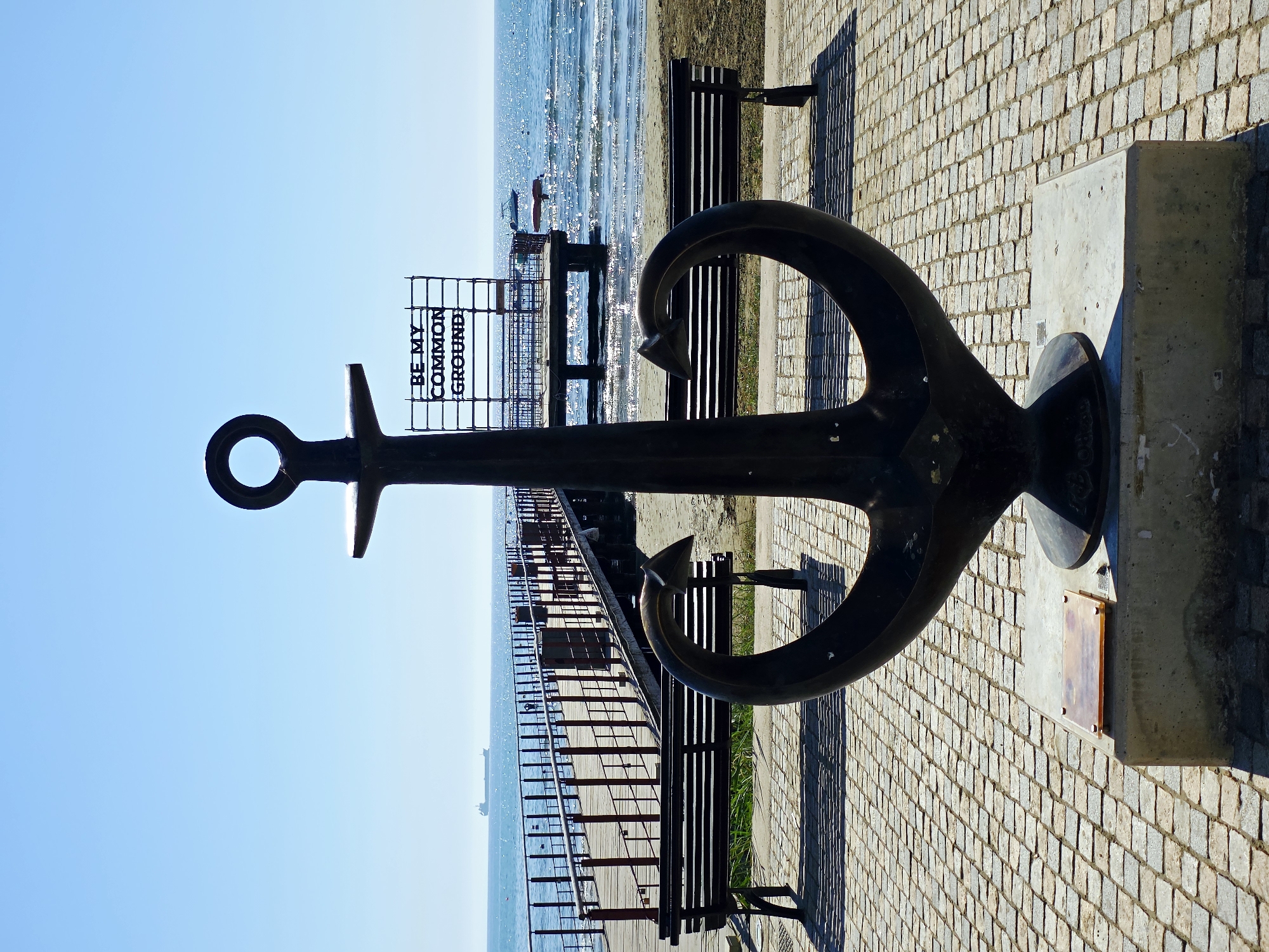 The image depicts a large, dark-colored anchor sculpture situated on a paved walkway in Larnaca, Cyprus, with the sea and a pier in the background. The anchor is the primary subject, positioned in the foreground and dominating the center of the frame. It rests on a concrete base, which has a small plaque on its front. Behind the anchor, a wooden pier extends into the blue sea. Benches are visible on either side of the walkway, casting long shadows in the sunlight. The sea is calm with gentle waves, and a large cargo ship is visible on the horizon. Above the pier, a sign with the text "BE MY COMMON GROUND" is mounted on a scaffolding-like structure. The sky is clear and blue. The environment suggests a seaside promenade, likely a tourist area due to the prominent sculpture and pier.