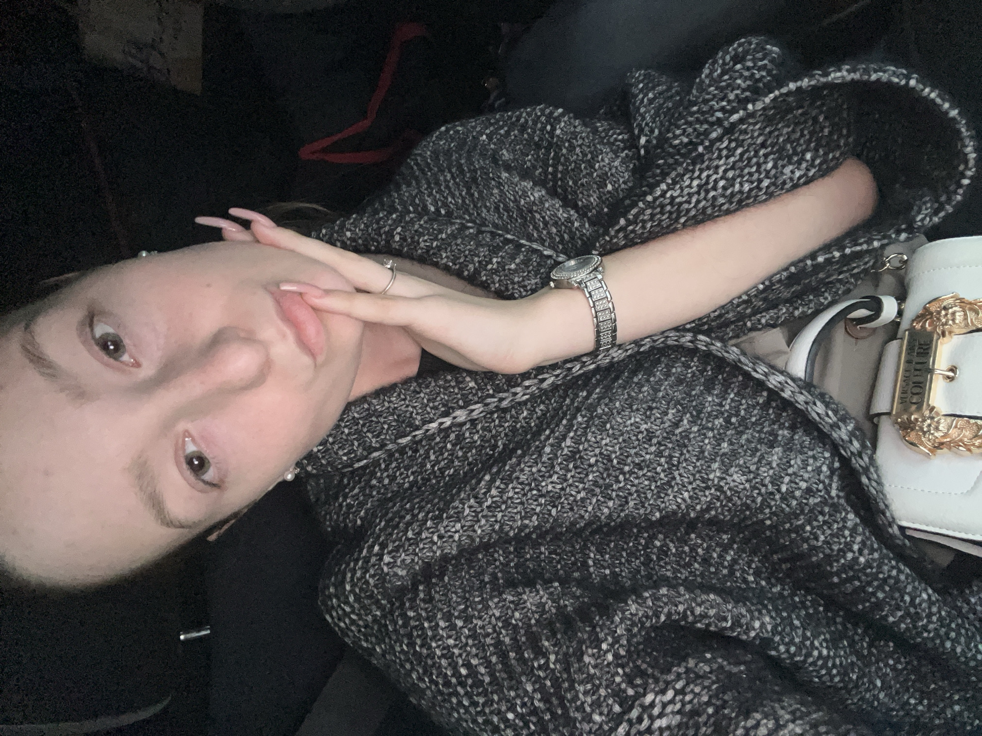 A fair-skinned person with light brown hair is indoors and is oriented in a way that their face is in the upper left quadrant of the frame. The person is wearing a silver wristwatch with a diamond-studded band on their left wrist and a ring on the ring finger of their left hand. They are wearing a gray and black patterned knit shawl or sweater, and partially visible is a white purse with a gold emblem.
The location is not possible to discern from the image provided. No specific landmarks or cultural items can be used to ascertain this to be the city of Varna, Bulgaria.
