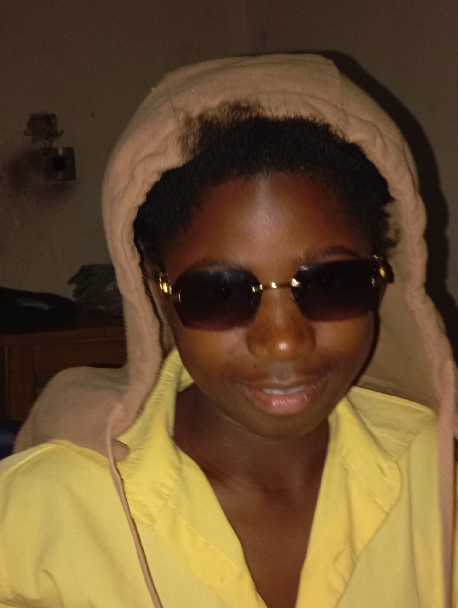 A person wearing a yellow collared shirt and sunglasses with gold accents is hooded in a beige material. The person is dark-skinned. The location appears to be indoors, given the lighting and the presence of a wooden structure and wall fixture in the background. It is not possible to confirm the location within the city of Jos, Nigeria based on the image.