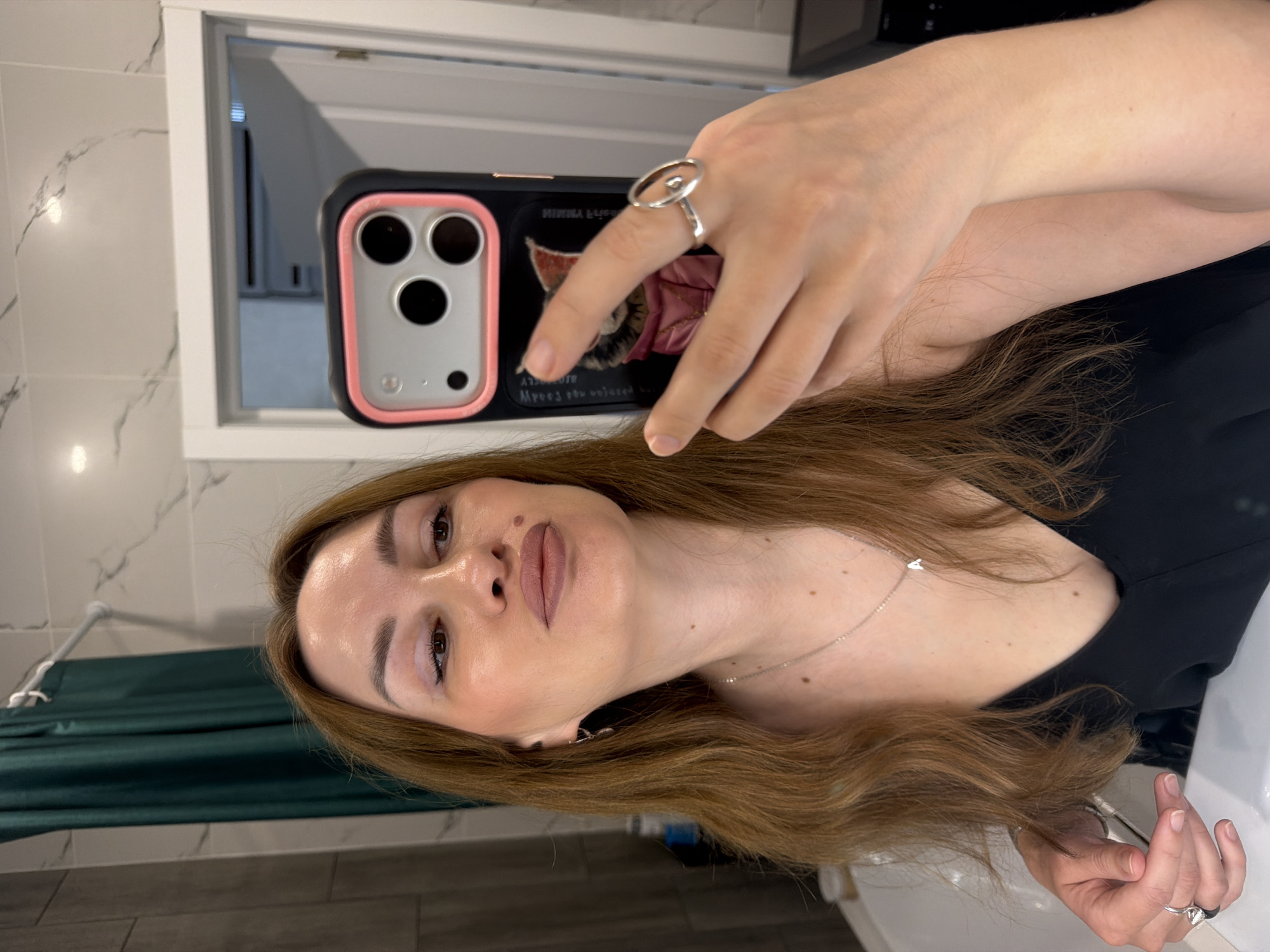 The image shows a woman taking a selfie in a bathroom. She has long, brown hair and is wearing a black top and jewelry, including rings and a necklace. The bathroom appears to be modern, with tiled walls and a green shower curtain. The woman is holding a phone in her right hand, and the phone case features a design of a cat. The lighting in the bathroom is soft, creating a warm atmosphere. The woman is looking at the phone's reflection in the mirror, with her lips slightly pursed.