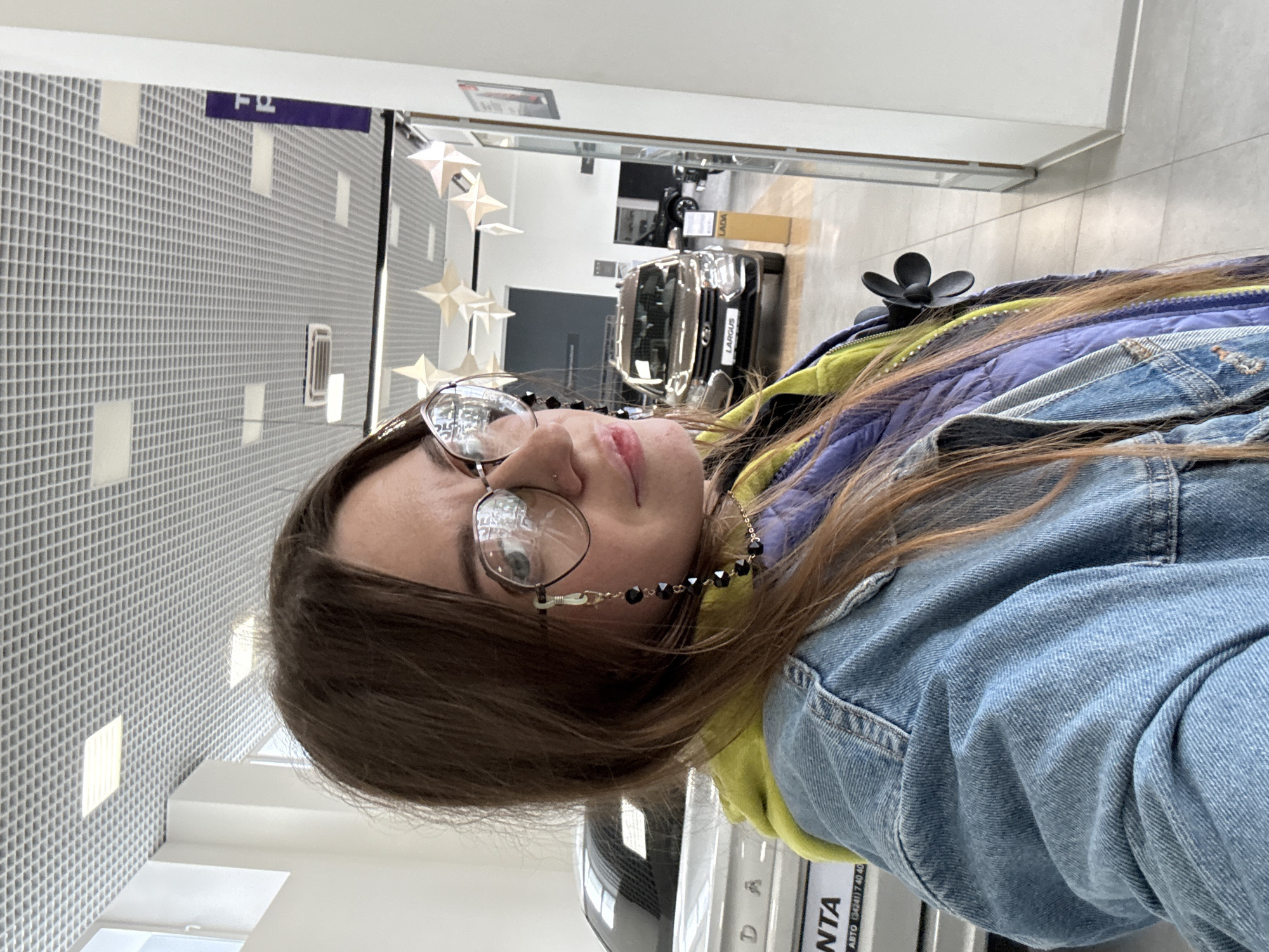 A woman with long brown hair and glasses is taking a selfie in a car dealership. She is wearing a denim jacket over a purple and yellow puffer vest. In the background, several cars are visible, including a Lada Largus. The dealership has a tiled floor and a suspended grid ceiling with fluorescent lighting. The woman's glasses have a beaded chain attached.