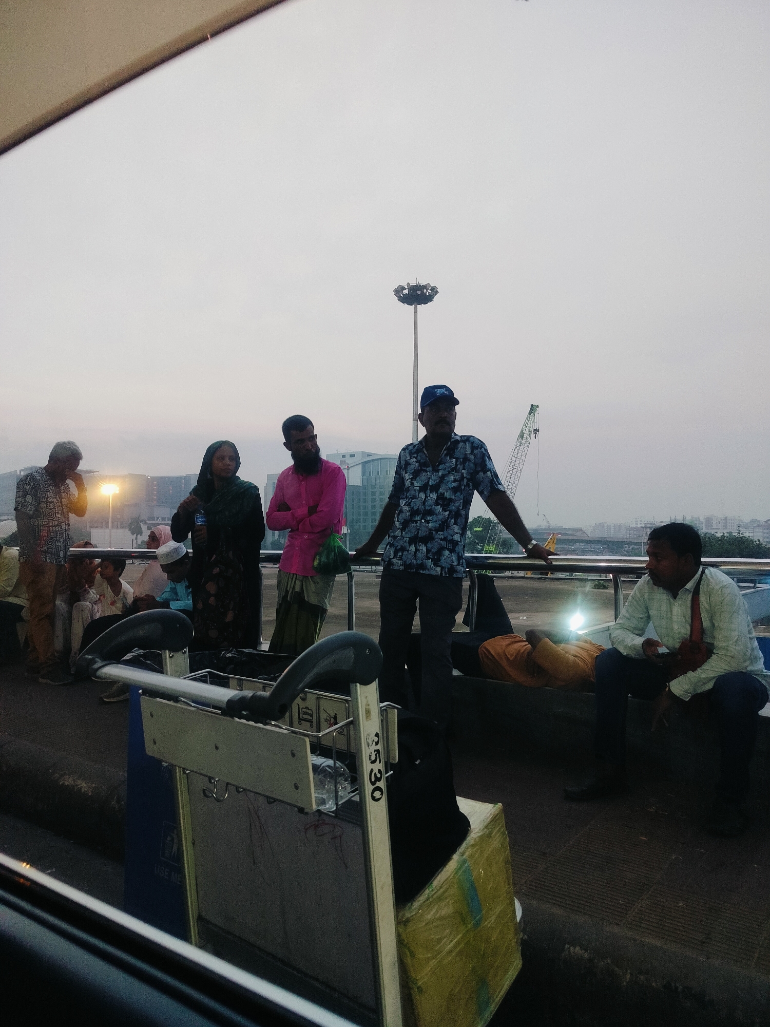 The image depicts a group of people gathered outdoors, likely in a public area of Dhaka, Bangladesh, possibly near an airport or transportation hub, given the presence of luggage carts. The sky is overcast, suggesting it might be dusk or dawn. Several individuals are standing, while others are seated or reclining on a bench. The people are dressed in a mix of traditional and modern attire. A prominent luggage cart is in the foreground. In the background, there are buildings, a tall light pole, and a construction crane. The overall scene appears to be a waiting area or a gathering point for travelers.