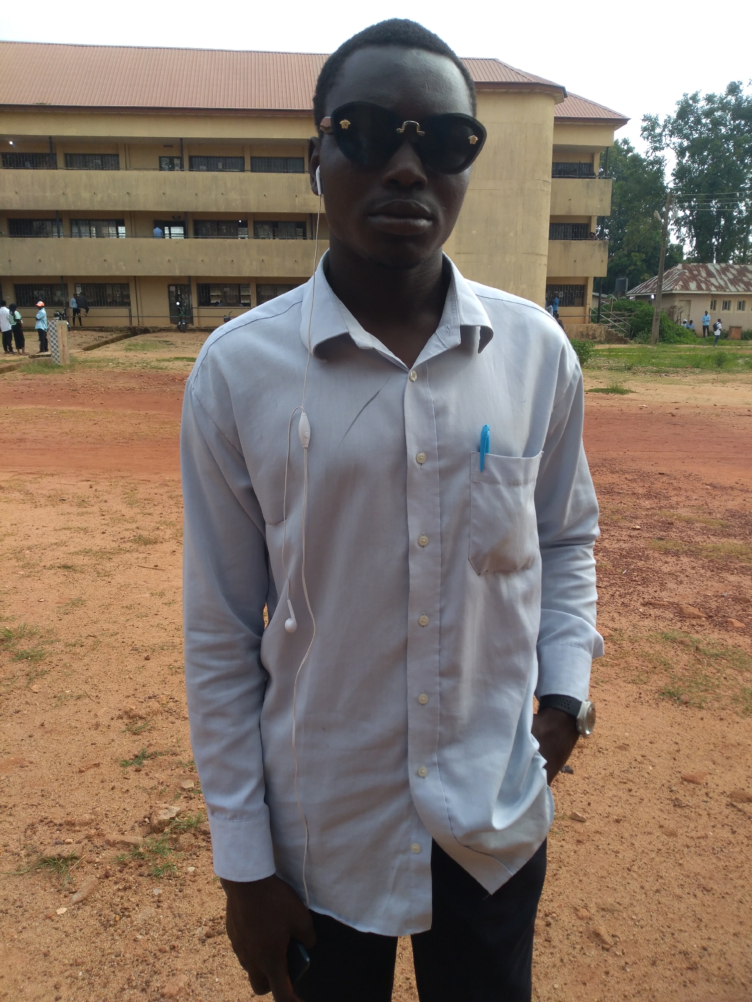 A young Black male stands in a dirt lot in front of a tan, multi-story building. He wears large black sunglasses, a light blue button-down shirt with a pen in the pocket, and black pants. White earbuds are visible in his ears and draped around his shirt. He has a watch on his left wrist and is holding a dark-colored smartphone in his right hand. In the background, the building appears to be a school or institutional facility with multiple windows and balconies. Other people are visible in the distance, near the building and on a path. The ground is covered in red dirt and sparse vegetation. The sky is overcast. The location is likely Jos, Nigeria, given the architecture and environment.