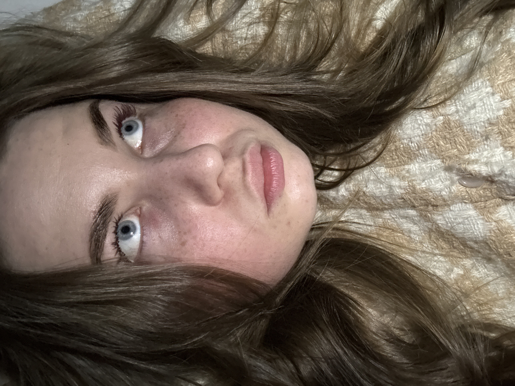 The image is a close-up, top-down portrait of a young person, likely female, lying down. The subject has fair skin with visible freckles across the nose and cheeks. Their eyes are a striking light blue, wide open, and gaze upward and slightly to the left, conveying a calm or pensive expression. Long, dark eyelashes, some showing a subtle reddish tint, frame the eyes, and the eyebrows are naturally well-defined and brown. The lips are full and have a soft pink hue.
Rich brown hair, appearing long and wavy, is spread out around the subject's head, blending into the background. The person is resting on a textured fabric, which features a distinct woven or checkered pattern in shades of cream, beige, and off-white. This background suggests an indoor, intimate setting, possibly a bed or sofa. The lighting is soft and even, highlighting the subject's features without harsh shadows.
There is no specific activity depicted beyond the person lying down and gazing. No location-specific cues are visible within the image itself to confirm the provided context of Chaikovsky, Russia. There is no visible text in the image.