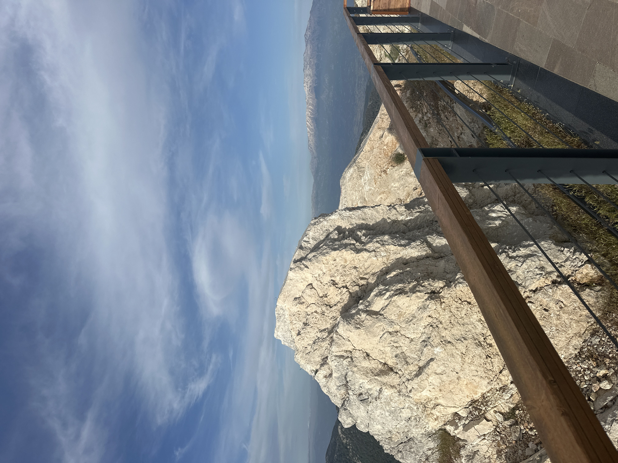The image shows a view from a cliffside perspective in Fethiye, Turkey. The sky is blue with scattered clouds. A cliff composed of light-colored, rough rock is prominent in the foreground. A metal and wooden railing runs along the edge of the cliff, suggesting a designated viewpoint or path. A mountainous landscape extends into the distance, fading with atmospheric haze. The overall impression is one of a high vantage point with a scenic view.