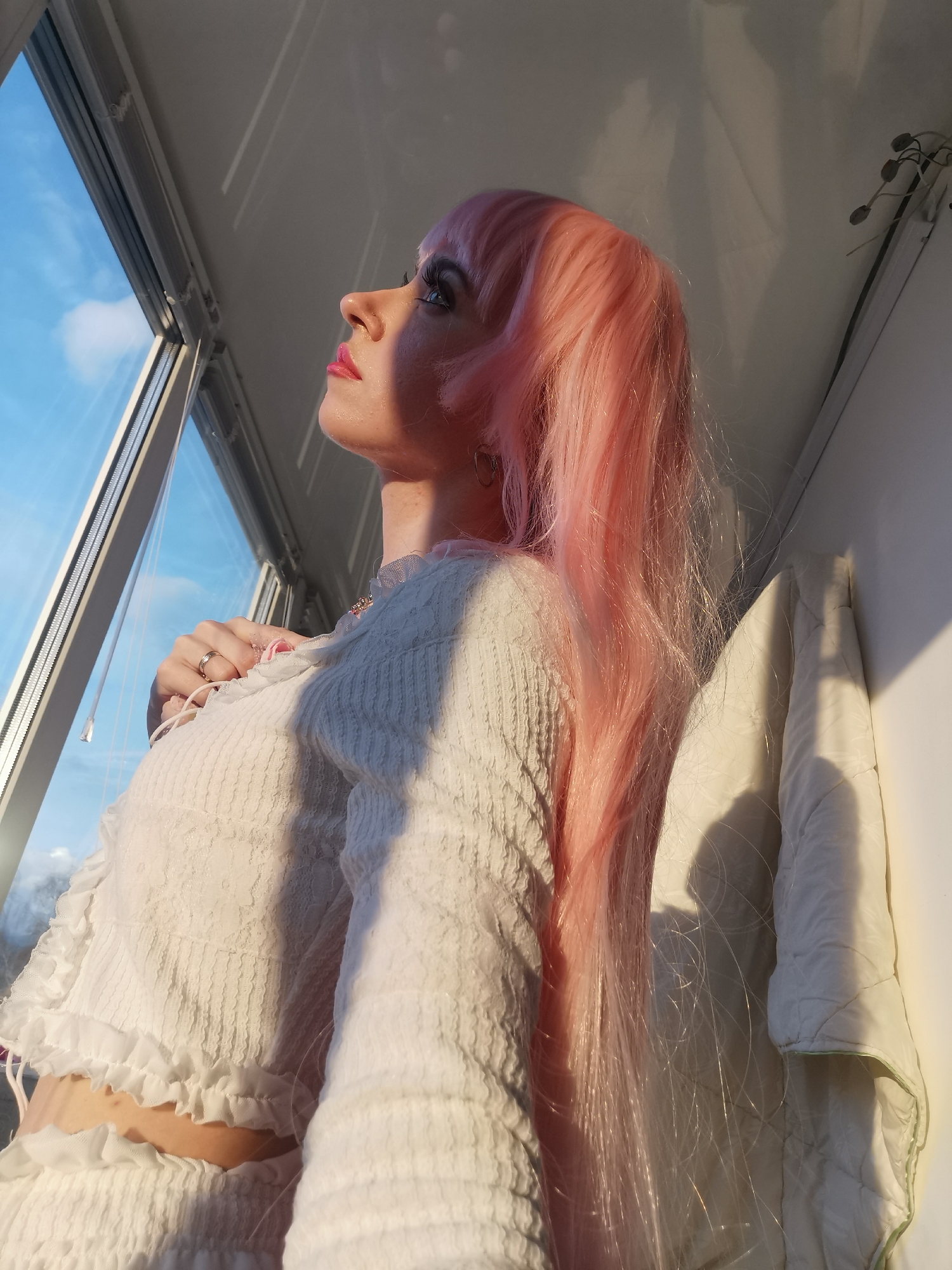 An indoor, eye-level shot captures a person, likely female, from the chest up, gazing out of a window. The subject has long, flowing pink hair and is wearing a white, textured top with ruffled trim and some lace detailing. A gold-toned ring is visible on their right ring finger, with that hand gently resting near their chest.
The person's profile is highlighted by warm, directional light, suggesting either early morning or late afternoon sun (golden hour), which casts soft shadows on their face and illuminates individual strands of hair. Their gaze is directed upwards and outwards, past the window frame, towards a clear blue sky with a few wispy white clouds.
The immediate surroundings include the white window frame and a white wall to the right of the person. A folded white, quilted item, possibly a blanket or curtain, hangs or rests against the wall, also touched by the warm sunlight. A dark, thin object resembling a light fixture or cable is visible on the upper right wall. The overall mood appears serene and contemplative, as the person looks towards the sky. There is no visible text.