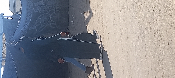A person wearing a long dark robe and jeans stands beside a makeshift structure covered with patterned fabric. Their head is also covered, and they appear to be holding an object in their hand. The ground is light-colored and rough, possibly concrete or a similar material. The scene is lit with bright sunlight, casting a sharp shadow of the person onto the ground. The image is rotated to the side.