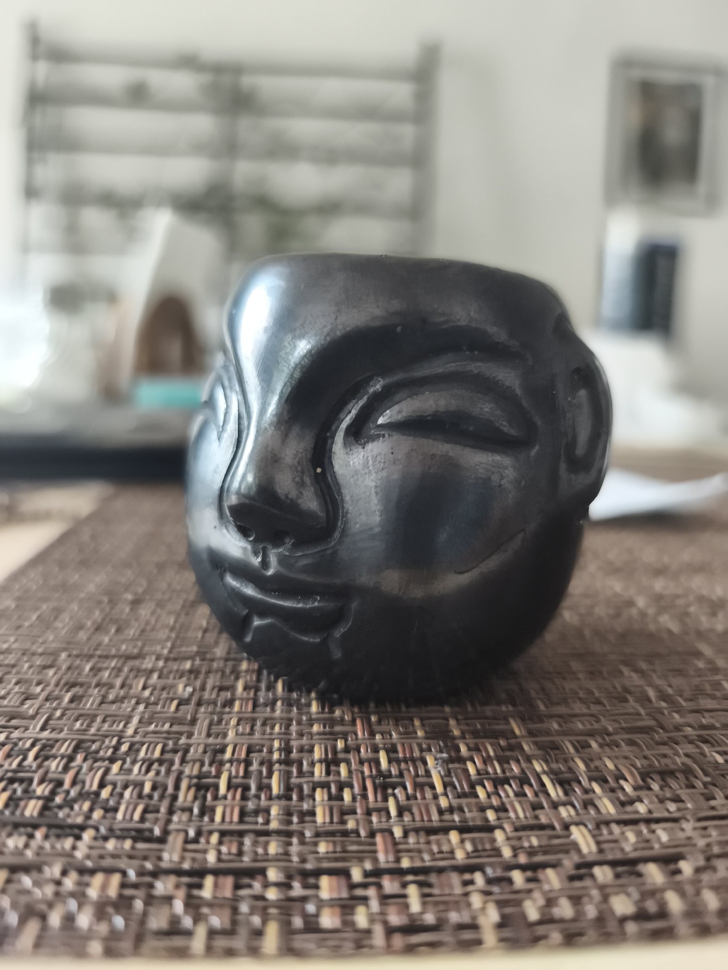 A close-up shot of a dark, glossy, sculpted head, resting on a woven brown placemat. The head has stylized facial features, including closed eyes, a nose, and a slightly smiling mouth. The background is blurred, but appears to show a window with blinds and other household objects on a table. The location cannot be determined based on visual information alone.