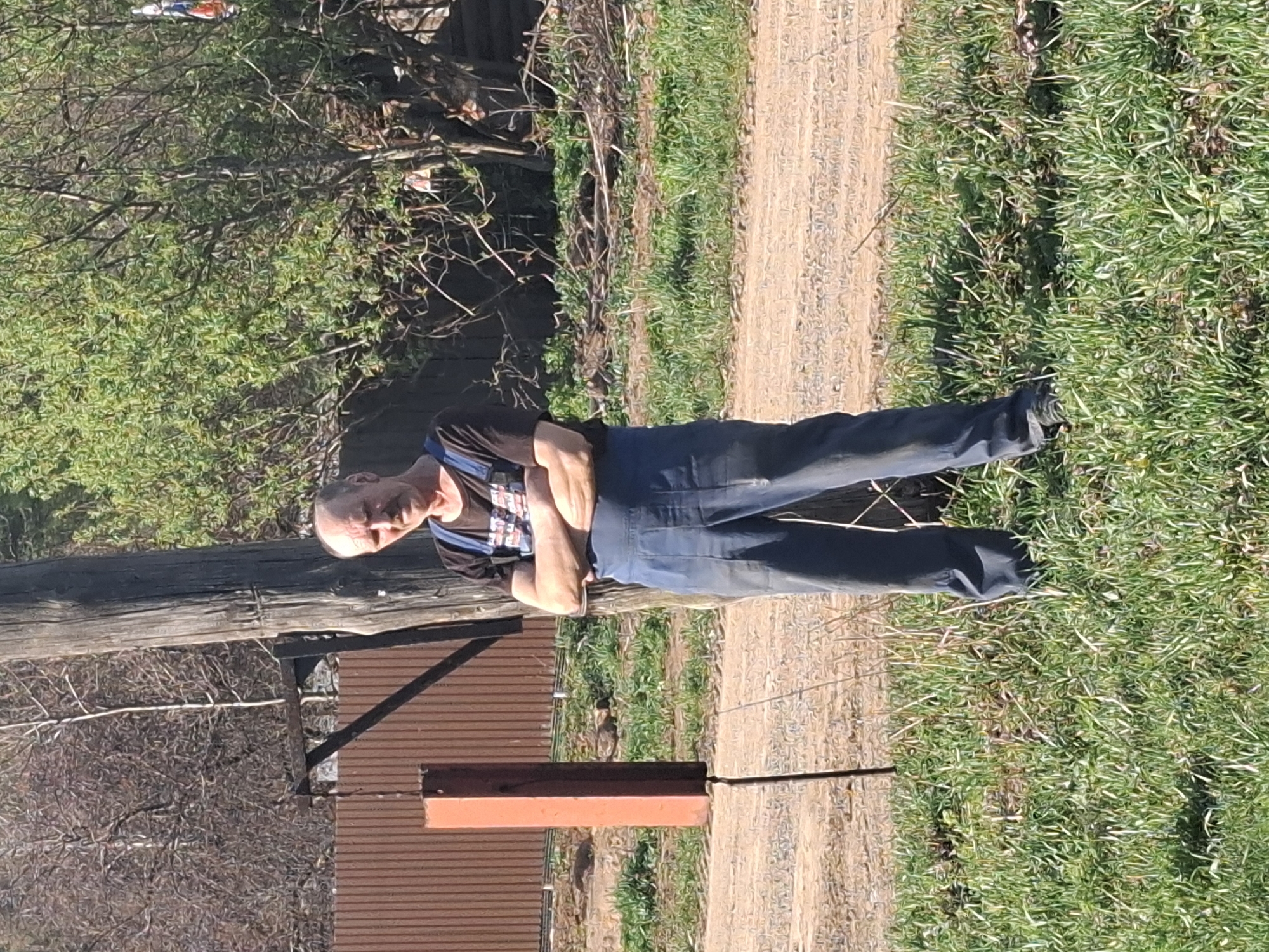 The image shows a man standing in an outdoor setting. He has a bald head and is wearing a black T-shirt underneath blue overalls. His arms are crossed. He is standing on a dirt and grass path. In the background, there is a building with a brown exterior and corrugated metal roofing. Additionally, there are trees and grass visible in the surrounding area. The location is possibly in Kamennyi Kliuch, Russia. A wooden post is visible to the left of the man.
