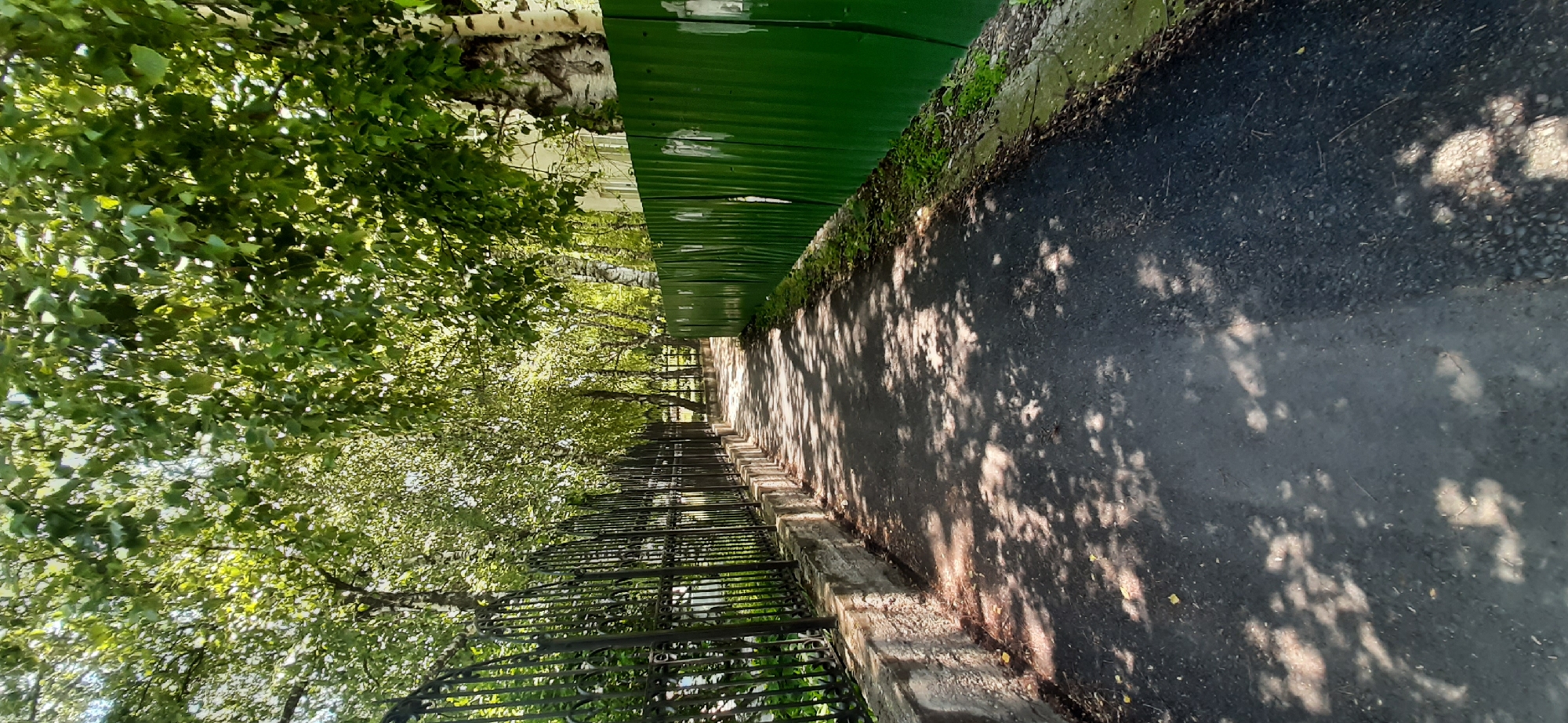 The image depicts a paved pathway bordered by a black metal fence on the left and a green corrugated metal fence on the right, likely within the city of Yessentukskaya, Russia. Overhead, the branches of trees with lush green leaves cast dappled shadows across the pathway. The pathway appears to be asphalt and shows signs of wear. The black fence is ornate with pointed finials and stands on a low stone wall. The green fence is taller and appears to be made of metal sheeting, with some white marks on its surface. The environment suggests a sunny day, possibly in a park or along a residential street. There are no people or animals visible in the image.