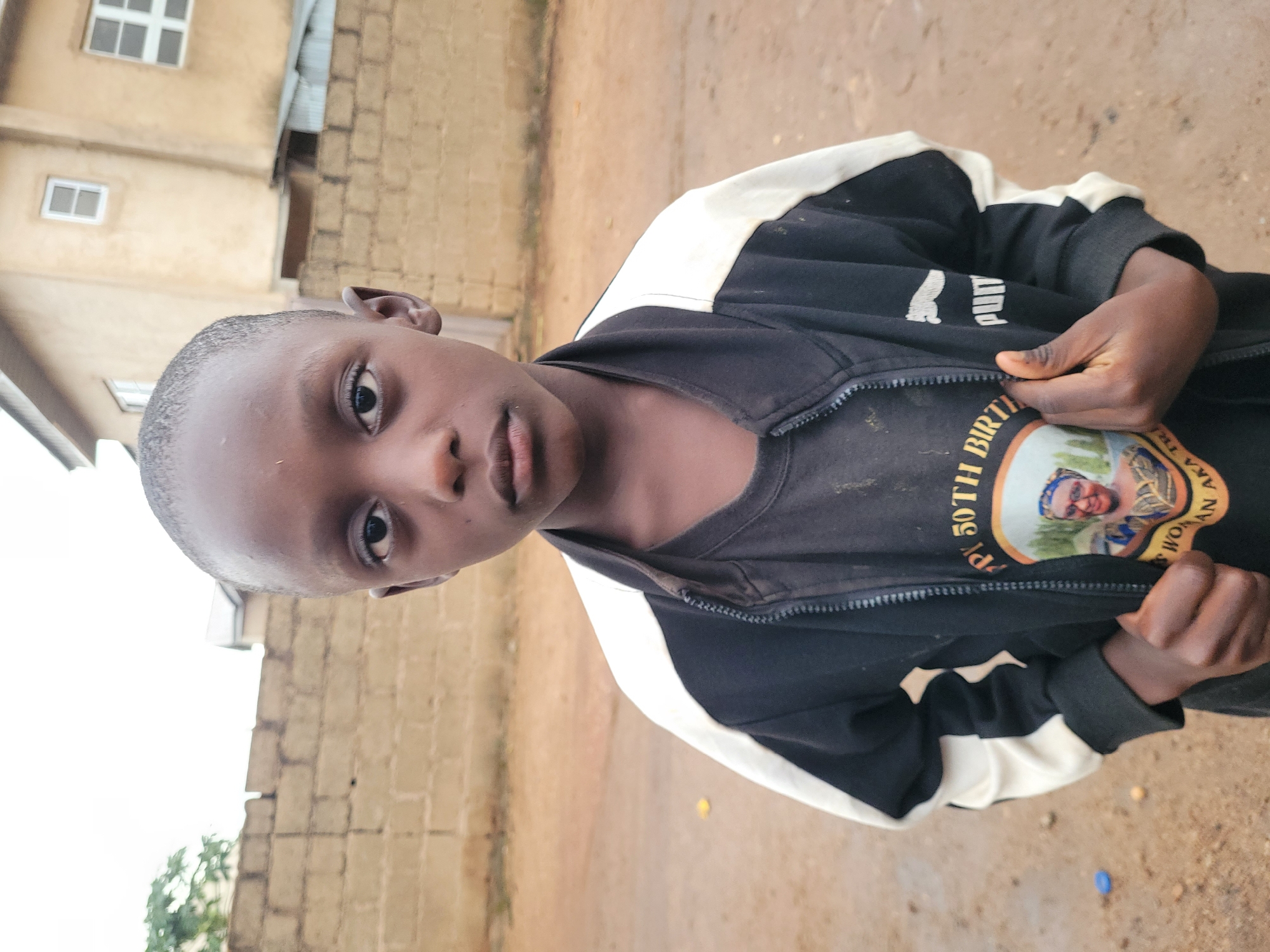 In Bukuru, Nigeria, a young boy stands outdoors, facing the camera directly. He has dark skin and a short haircut. He is wearing a black t-shirt with a circular graphic featuring a woman's face and text that partially reads "HAPPY 50TH BIRTH... WOMAN AKA TRI...". Over the t-shirt, he wears a black and white zip-up jacket. The background consists of a light brown, dirt ground, a brick wall, and a building with windows. The sky is overcast. The boy is holding the zipper of his jacket with his right hand.