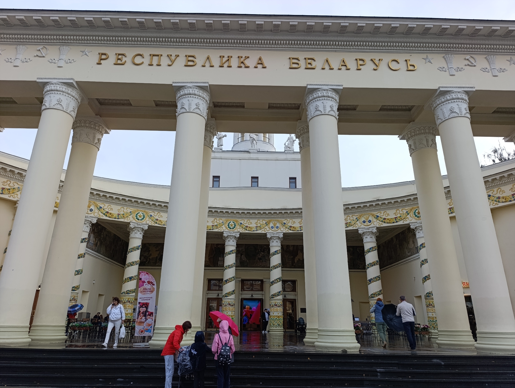 The image depicts the exterior of the pavilion of the Republic of Belarus at VDNKh (Exhibition of Achievements of National Economy) in Moscow, Russia. The grand facade features a semicircular colonnade with massive, ornate white columns. Above the columns, a frieze displays golden Cyrillic lettering that reads "РЕСПУБЛИКА БЕЛАРУСЬ" (Republic of Belarus), flanked by decorative elements including stars, wheat sheaves, and the hammer and sickle symbol. The building itself is light-colored with classical architectural details. In the foreground, several people are visible on dark steps leading up to the pavilion. Some are walking away from the camera, while others are looking towards the entrance. One person is holding a bright pink umbrella. The ground appears wet, suggesting recent rain. Banners and posters are visible near the entrance, promoting various exhibits or services. The overall scene conveys a sense of architectural grandeur and public gathering at an exhibition space.