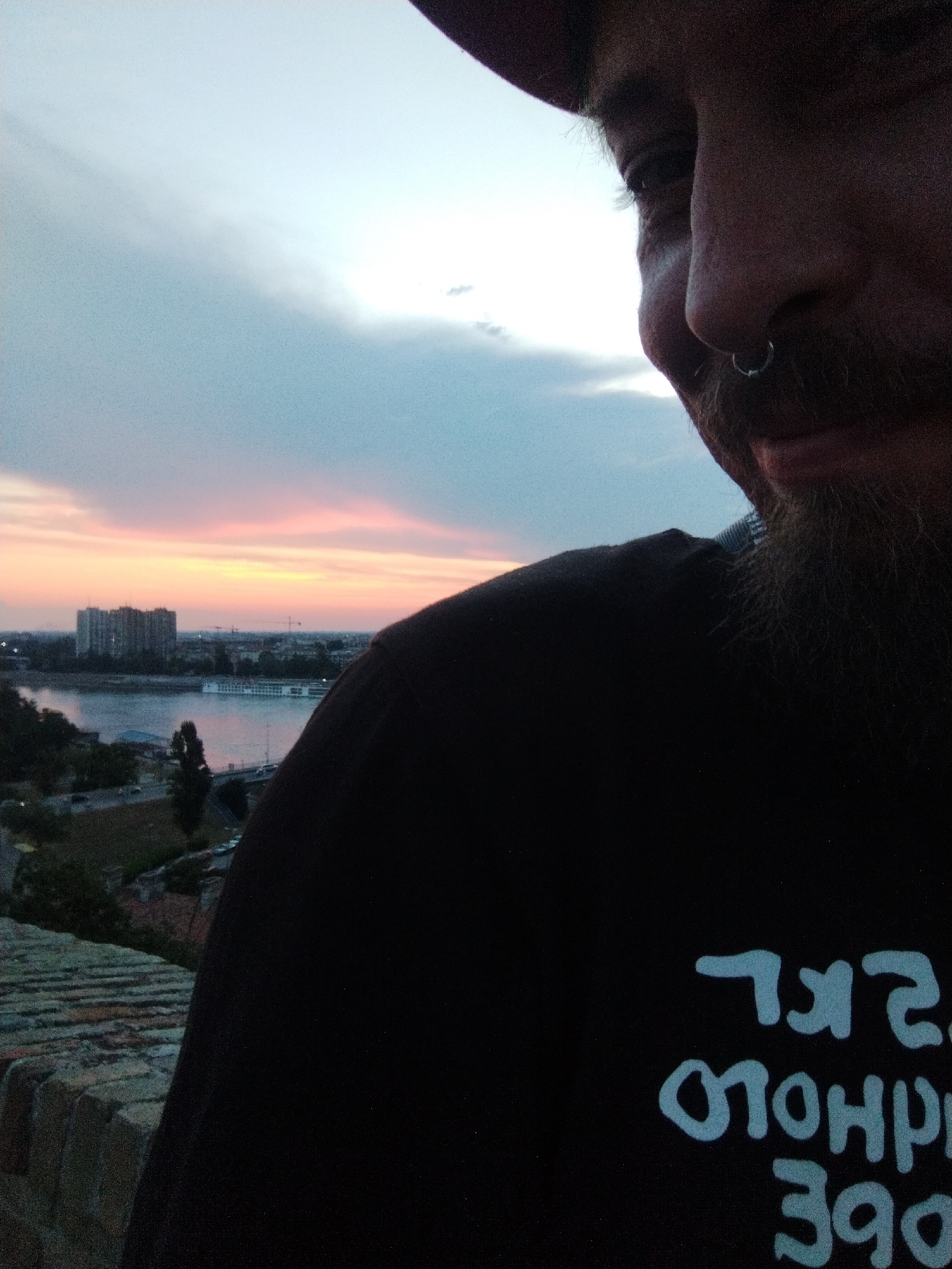 The image is a close-up, low-angle selfie taken outdoors at dusk, likely from the Petrovaradin Fortress in Petrovaradin, Serbia, overlooking the Danube River and the city of Novi Sad. In the foreground, a person's face is partially visible on the right side, with a beard and a nose ring. They are wearing a black t-shirt with light blue text printed on it. The text is partially obscured and appears to be in a Cyrillic or similar script, with some letters appearing reversed. The background features a hazy sky with a pink and orange sunset, a wide river, buildings on the opposite bank, and some trees. A brick wall or parapet is visible in the lower left corner, suggesting the photo was taken from an elevated position.