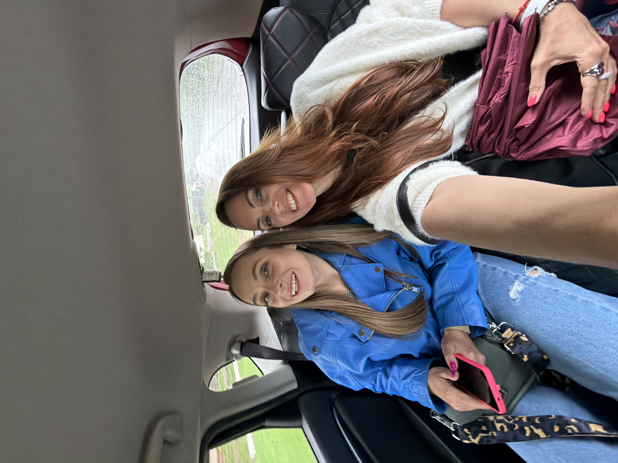 Two women are sitting in the back of a car. The woman closest to the camera is wearing a blue jacket and light wash jeans. She is holding a cell phone in her hand. The woman behind her is wearing a white sweater. It is raining outside. The image does not contain enough details to determine the location in Birobidzhan, Russia.