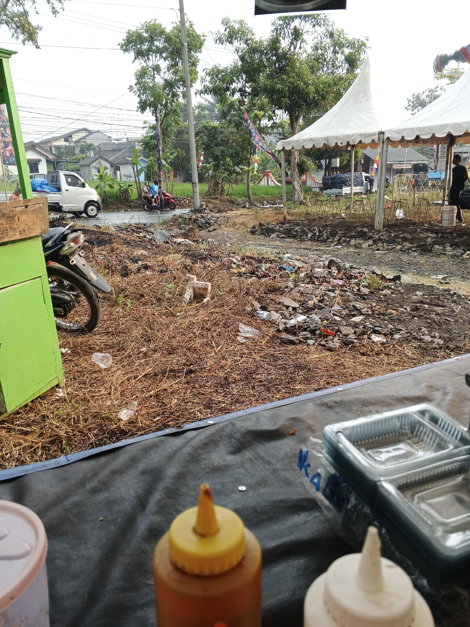 The image depicts an outdoor scene in Sukabumi, Indonesia, characterized by a cluttered and somewhat unkempt environment. In the foreground, a black tarp covers a surface, upon which several items are placed, including a stack of clear plastic food containers, a bottle with a yellow cap, and another bottle with a white cap. To the left, a portion of a bright green food stall or cart is visible, along with the rear wheel and fender of a motorcycle.
The middle ground of the image reveals a patch of dry, brown vegetation interspersed with debris, including rocks, plastic bottles, and other trash. Beyond this, a paved road is present, on which a white pickup truck and a red motorcycle with two occupants are traveling. Several trees with green foliage dot the landscape.
In the background, residential buildings with tiled roofs are visible, suggesting a semi-urban or suburban setting. Two white tents with pointed roofs stand on the right side of the image, with a person standing near one of them. A utility pole with overhead wires is also prominent. The sky is overcast, indicating a cloudy day. The overall impression is one of a temporary outdoor market or event setup that has been left with considerable debris.
