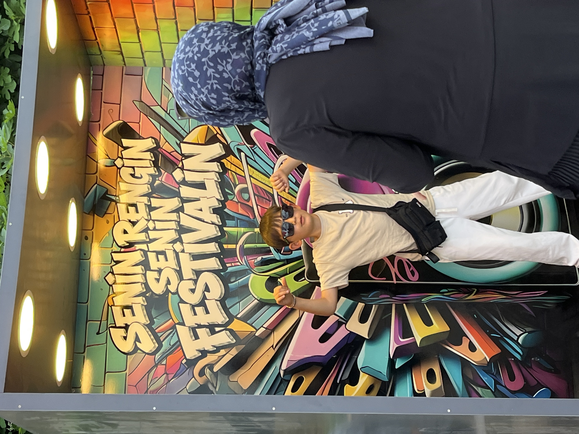 The image shows an individual wearing sunglasses and a beige t-shirt, posing in front of a "SENİN RENK SENİN FESTİVALİN" (Your Color, Your Festival) themed installation. The installation appears to be a photo booth or art display, with graffiti-style lettering and colorful abstract designs. The person is wearing white pants and a black shoulder bag, giving a thumbs-up. Above the young man's head is a woman in a hijab. The setting appears to be outdoors. Alanya, Turkey can be recognized due to the text of the mural on the wall.