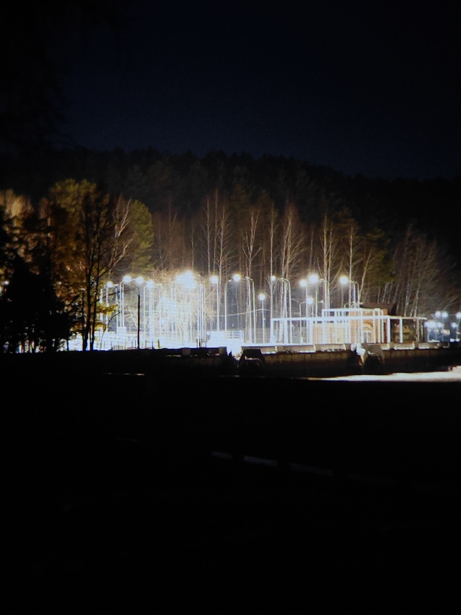 The image depicts a nighttime scene in Chaikovsky, Russia. In the middle ground, a fenced-in area with numerous lampposts illuminating the space is visible. Within the fenced area, there is a structure with white supports and what appears to be a covered area. Beyond the fenced area, a densely wooded area is present. In the foreground, there is a dark, undefined area. The overall atmosphere is dark, with the only sources of light emanating from the lamps within the fenced area.