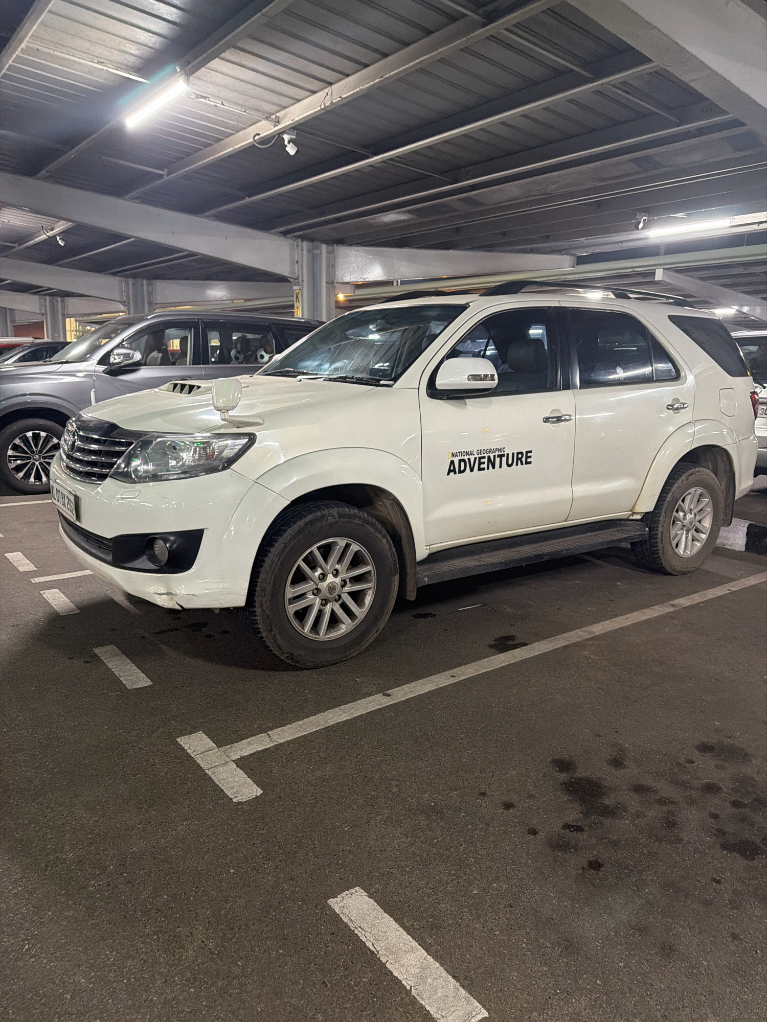 A white Toyota Fortuner is parked in a covered parking lot in Kochi, India. The vehicle has a "NATIONAL GEOGRAPHIC ADVENTURE" sticker on the driver's side door. The parking lot has a corrugated metal roof and fluorescent lighting. The ground is asphalt and has white parking space markings. Another car, a grey SUV, is parked to the left of the Fortuner. The image appears to have been taken at night or in low light conditions.