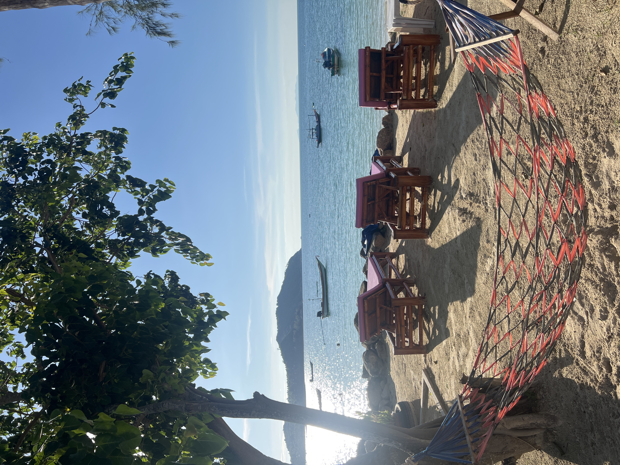 The image depicts a serene beach scene on Koh Phangan, Thailand, under a clear blue sky. In the foreground, a hammock with red and blue netting is strung between a tree and a wooden stand. The sandy beach is dotted with four wooden lounge chairs with pink cushions, facing the turquoise ocean. Several boats are visible in the water, with a larger island in the background. Sunlight glints off the water, creating a shimmering effect. The overall atmosphere is peaceful and inviting, characteristic of a tropical vacation destination.