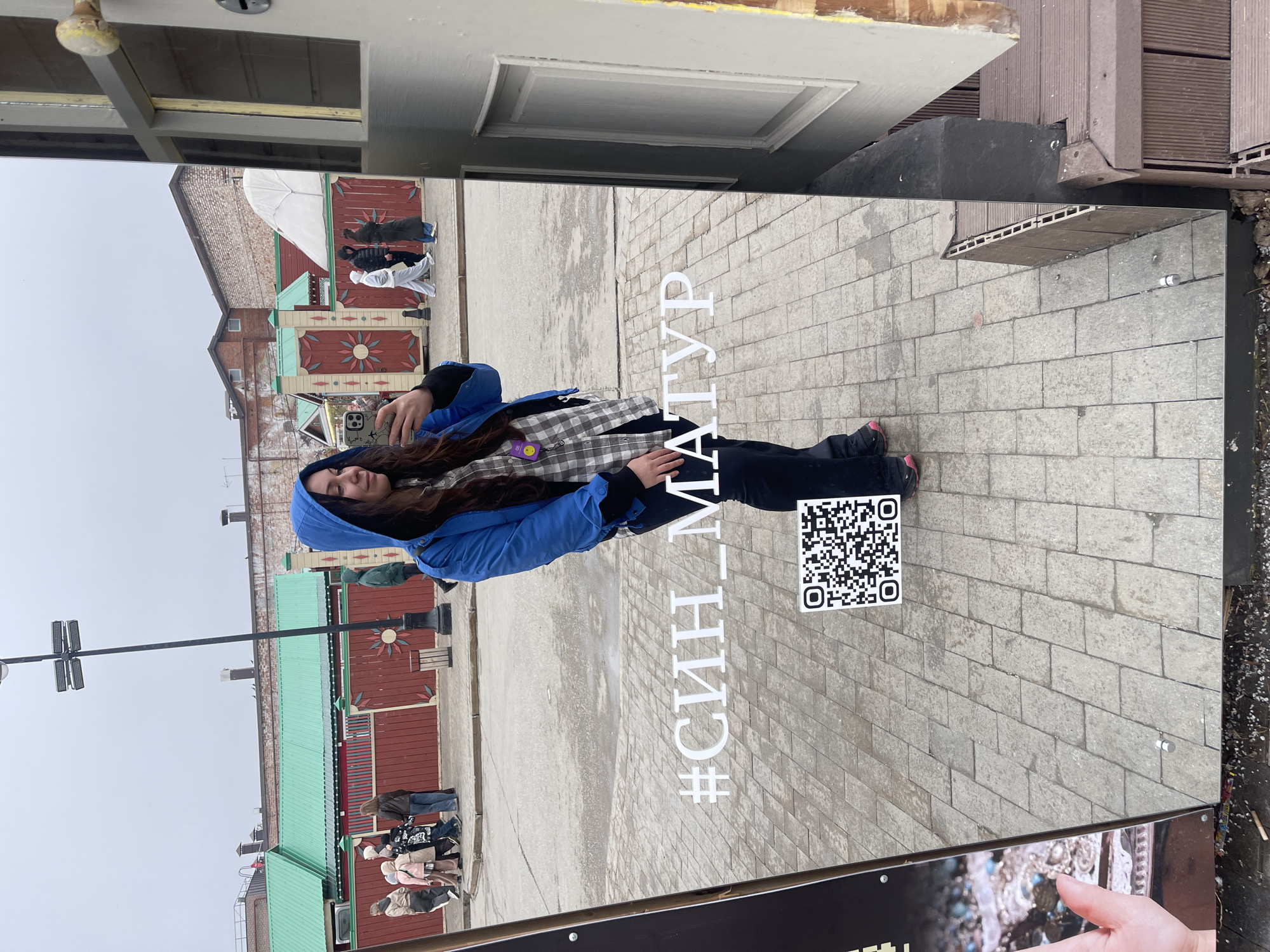 A long-haired woman in a blue hooded jacket and gray checkered shirt is visible in the reflection of a sign that has the hashtag "#СИН_МАТУР" and a QR code on it. The sign is positioned in an urban environment, likely the city of Kazan, Russia, as suggested by the Cyrillic text. In the background, there are red buildings with distinctive painted decorations, typical of Tatar architecture. Pedestrians are also visible in the background, suggesting a public space.