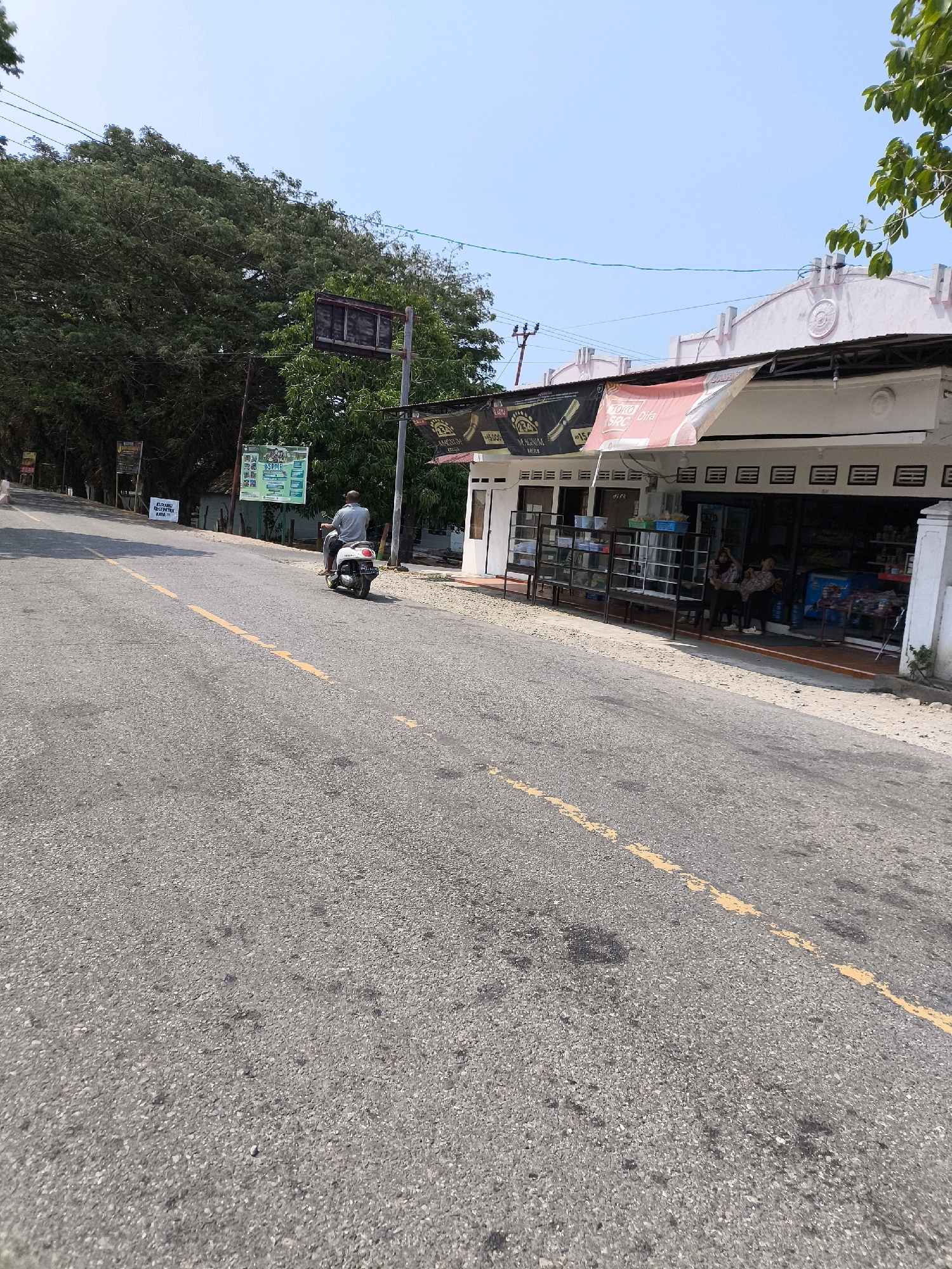 The image captures a street scene in an outdoor environment, likely in a city or town in Indonesia, given the signage visible. The foreground is dominated by a paved road with a yellow dashed line running down its center, indicating a two-way traffic lane. The road surface appears somewhat worn and speckled with dark patches.
To the right of the road, a low-rise building with a white facade stretches along the street. The building has an awning extending outwards, displaying advertisements. The awning has a red and white banner on the left side with Indonesian text and a black banner on the right side, also with Indonesian text and images of what appear to be cigarettes or similar products. Beneath the awning, a row of open-fronted shops or stalls is visible. Some of these stalls have glass display cases. Inside the shops, shelves are stocked with various items, and a few people are standing or sitting. Two women are seen near the entrance of one of the shops.
In the middle ground, a person is riding a white scooter on the road, moving away from the viewer. The rider is wearing a light-colored shirt and shorts. To the left of the road, there is a dense area of trees with lush green foliage. A traffic light signal is mounted on a pole near the trees. In front of the trees, there are several signs, including a green and white sign with text in Indonesian.
The sky above is clear and blue with no visible clouds, suggesting a sunny day. The overall scene depicts a typical roadside commercial area with a mix of residential or business buildings and natural vegetation.