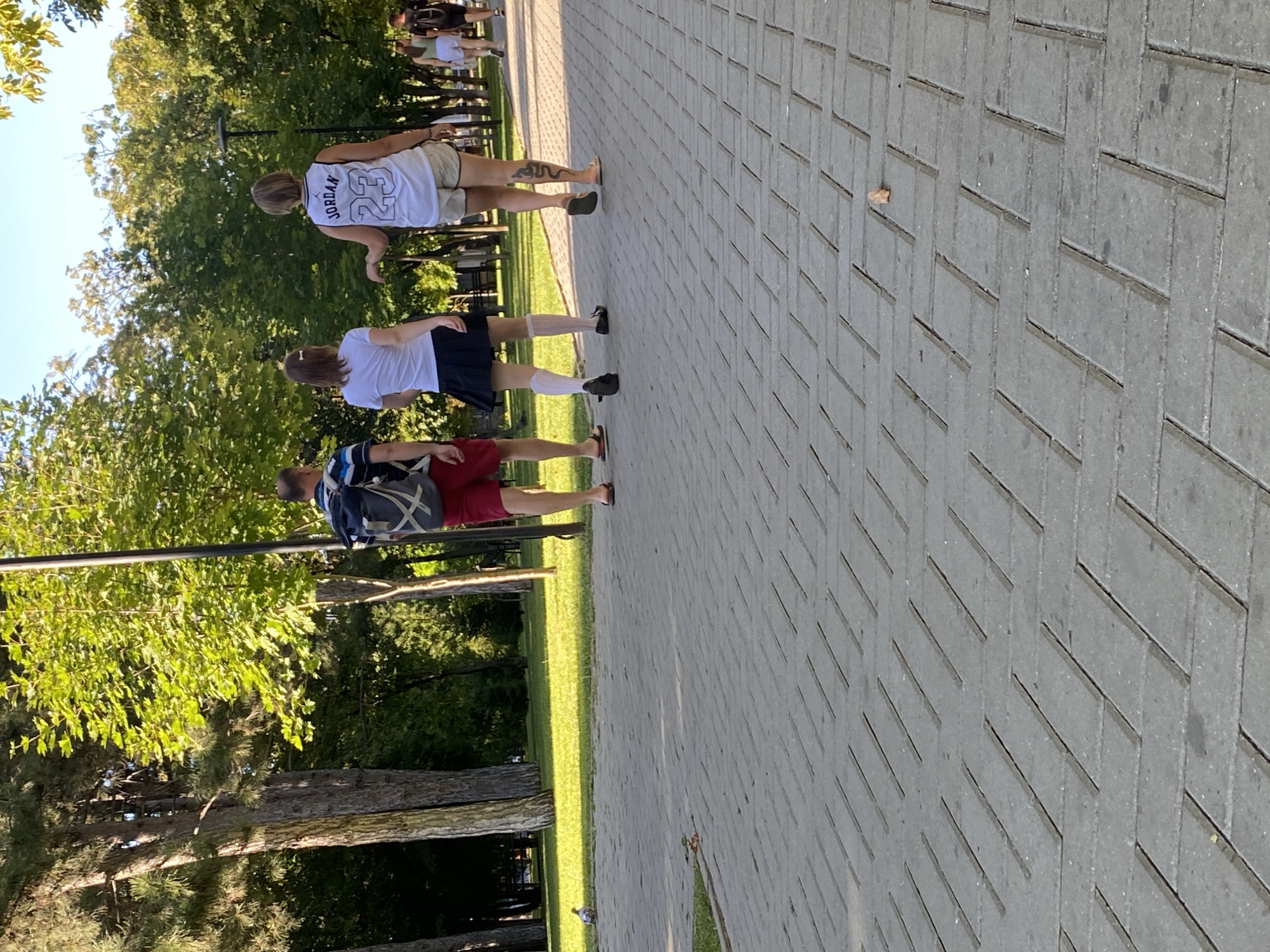 A group of people walk on a paved walkway lined with trees and grass in what appears to be a park. The foreground shows a close-up of the gray, rectangular pavers that make up the walkway. In the middle ground, a man wearing red shorts and a backpack walks away from the camera. To his right, a young woman in a white top and black skirt walks ahead. Another woman in a white tank top with "JORDAN 23" on it and light-colored shorts walks to the right of the young woman, with a tattoo visible on her calf. In the background, several other individuals are walking further down the path. The overall scene suggests a casual outing in a public park on a sunny day. The specific location in Chișinău, Moldova is not discernible from the visual information provided.