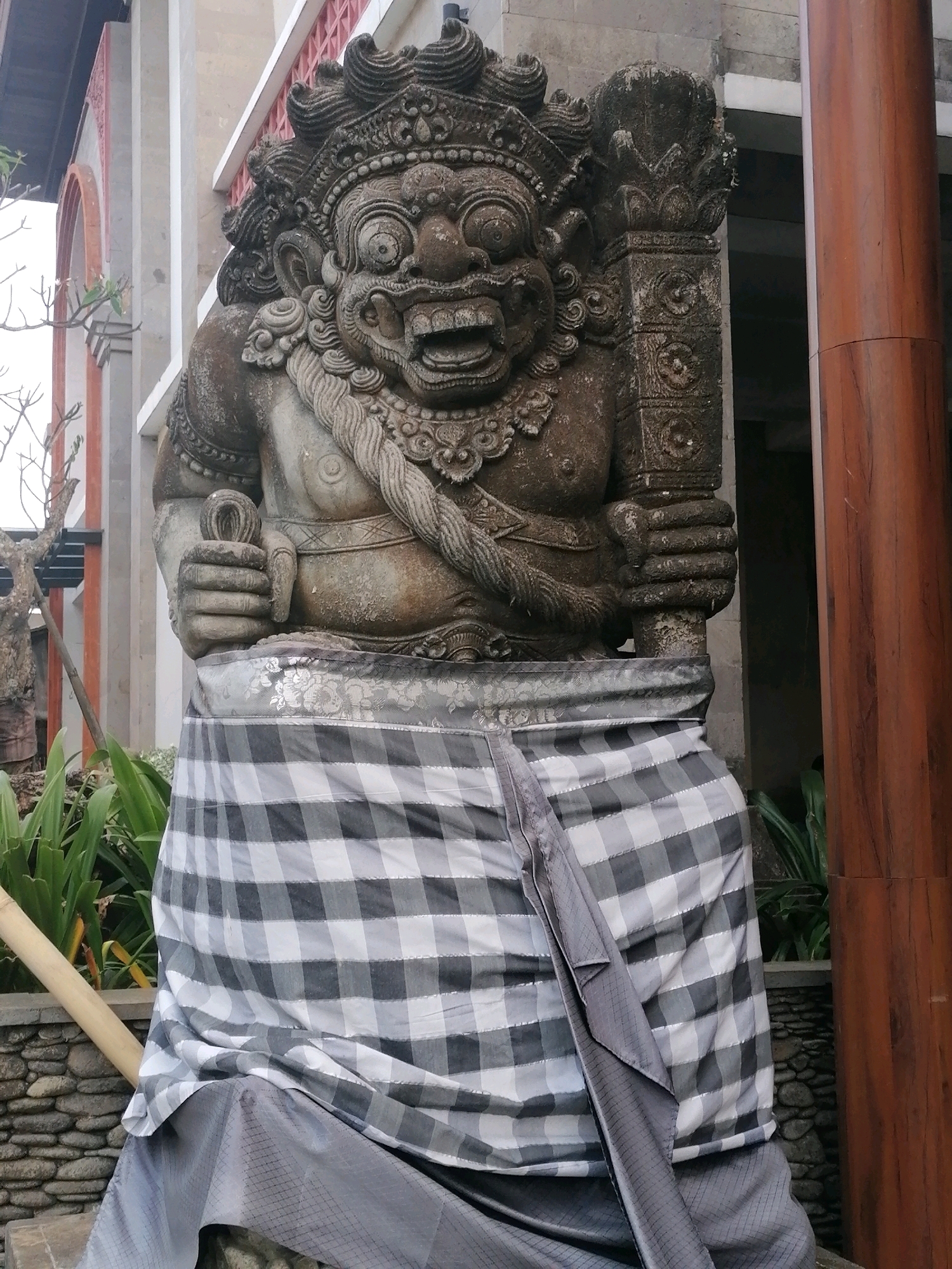 The image depicts a stone statue of a fierce deity, possibly a Balinese Barong or Rangda, with a menacing expression, elaborate headdress, and bared teeth. The statue is clad in a traditional Balinese checkered sarong. It stands outdoors in front of a building with architectural features that suggest a temple or a traditional Balinese establishment. The background includes greenery, stone pathways, and a large wooden pillar, consistent with the tropical environment of Ubud District, Indonesia. The statue appears to be a decorative element in a public or semi-public space.
