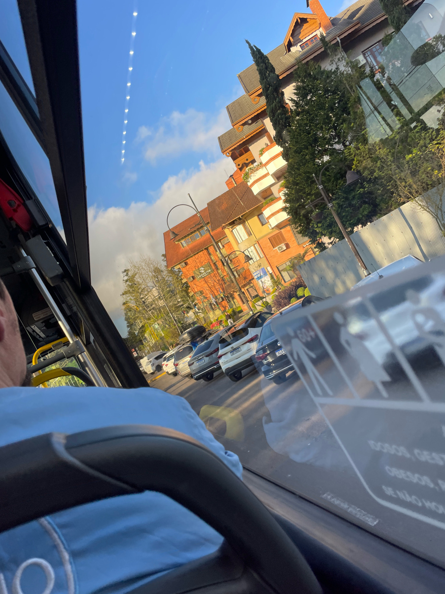 The image shows a view from inside a bus, looking out at a street scene in Gramado, Brazil. The bus has a black interior with a blue seat and yellow grab bar visible. The passenger is wearing a light blue shirt. Outside, a row of cars is parked along the street. The buildings in the background are multi-story structures with orange and white facades, some with a rustic architectural style. Trees are interspersed among the buildings. The sky is clear blue with some scattered white clouds. The light suggests it is daytime, possibly late afternoon. Reflections on the bus window show part of the interior and the exterior scene. A sign with silhouettes of people and text, possibly indicating priority seating, is visible on the window.