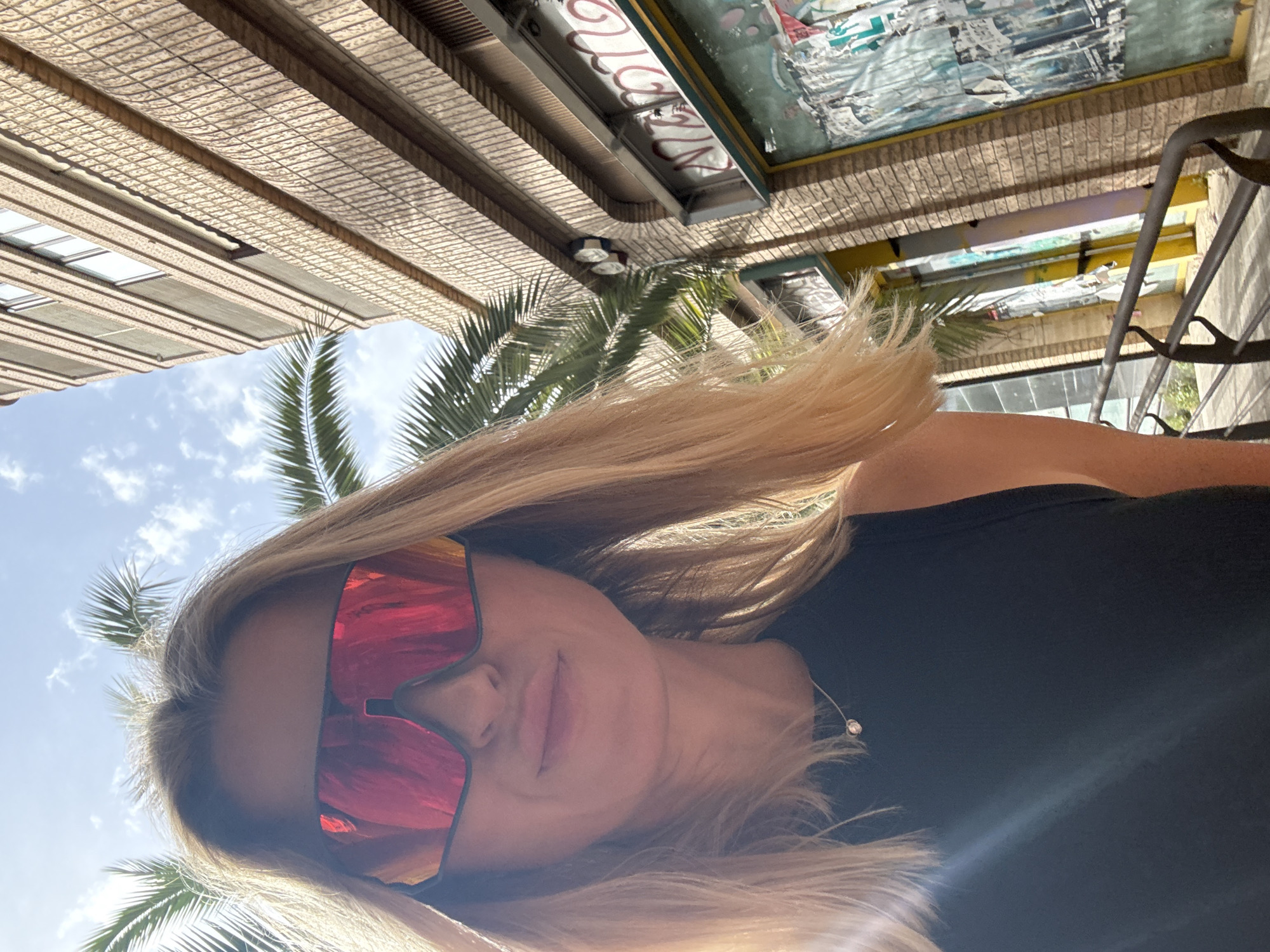 A medium shot, outdoors, features a woman with blonde hair and large, reflective red sunglasses. She wears a sleeveless black top with a delicate silver necklace. Behind her, a palm tree with vibrant green fronds stands against a bright blue sky with scattered clouds. To the right, a tan brick building with horizontal textured bands and multiple windows rises upwards. The lower part of the building has a storefront with graffiti tags and peeling posters on a green-tinted glass facade. A dark metal railing runs horizontally in the foreground. The scene appears to be in a Mediterranean climate, possibly Valencia, Spain, given the architecture and vegetation.
