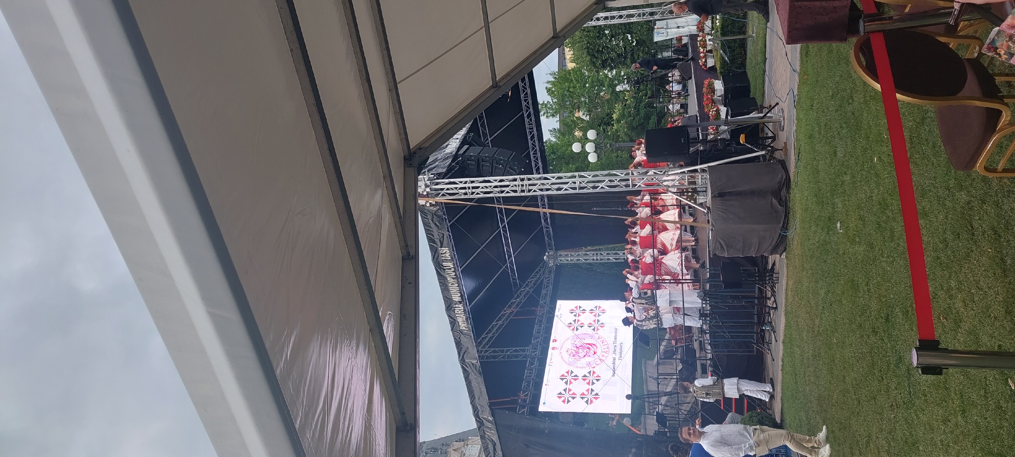 The image captures an outdoor event, likely a performance, under a cloudy sky. A large stage with a black backdrop is set up, featuring a prominent LED screen displaying a logo and text. The text on the screen indicates "PRIMARIA MUNICIPIULUI IASI," suggesting the event is sponsored or hosted by the City Hall of Iași, Romania.
On the stage, a group of performers, dressed in traditional folk costumes with intricate embroidery, are engaged in a dance. The costumes are predominantly white with red and black decorative elements. Some performers wear vests over white shirts, and the women wear full skirts. They are positioned in a circle or semi-circle, holding hands.
In the foreground, under a large white tent structure, there is a red velvet rope barrier separating the audience area from the performance space. Several chairs are visible behind the barrier. The ground is covered in green grass.
To the left of the stage, a man in a white shirt and beige pants stands on a small platform, looking towards the performance. To the right of the stage, a cameraman is filming the event, and a person in dark clothing stands near some sound equipment.
The overall impression is of a cultural event or festival taking place in a public space in Iași.